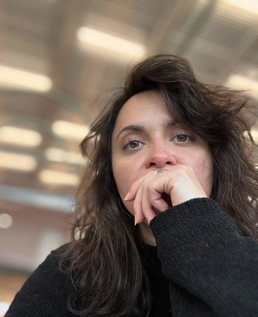 Close-up of a woman with dark, wavy hair, wearing a black sweater, in an indoor setting with blurred ceiling lights.