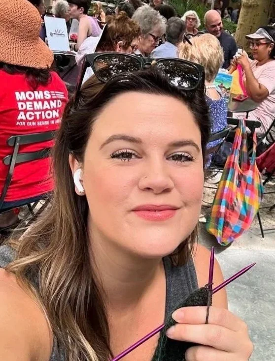 A young woman taking a selfie at an outdoor event, surrounded by people sitting at tables. She has brown hair, sunglasses on her head, and is holding knitting needles with yarn.
