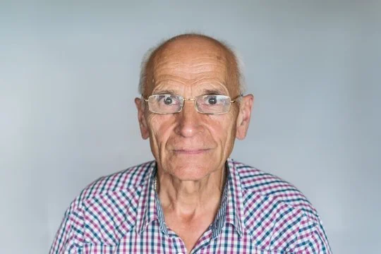 Close-up of an elderly man with glasses wearing a checkered shirt, looking at the camera against a plain gray background.