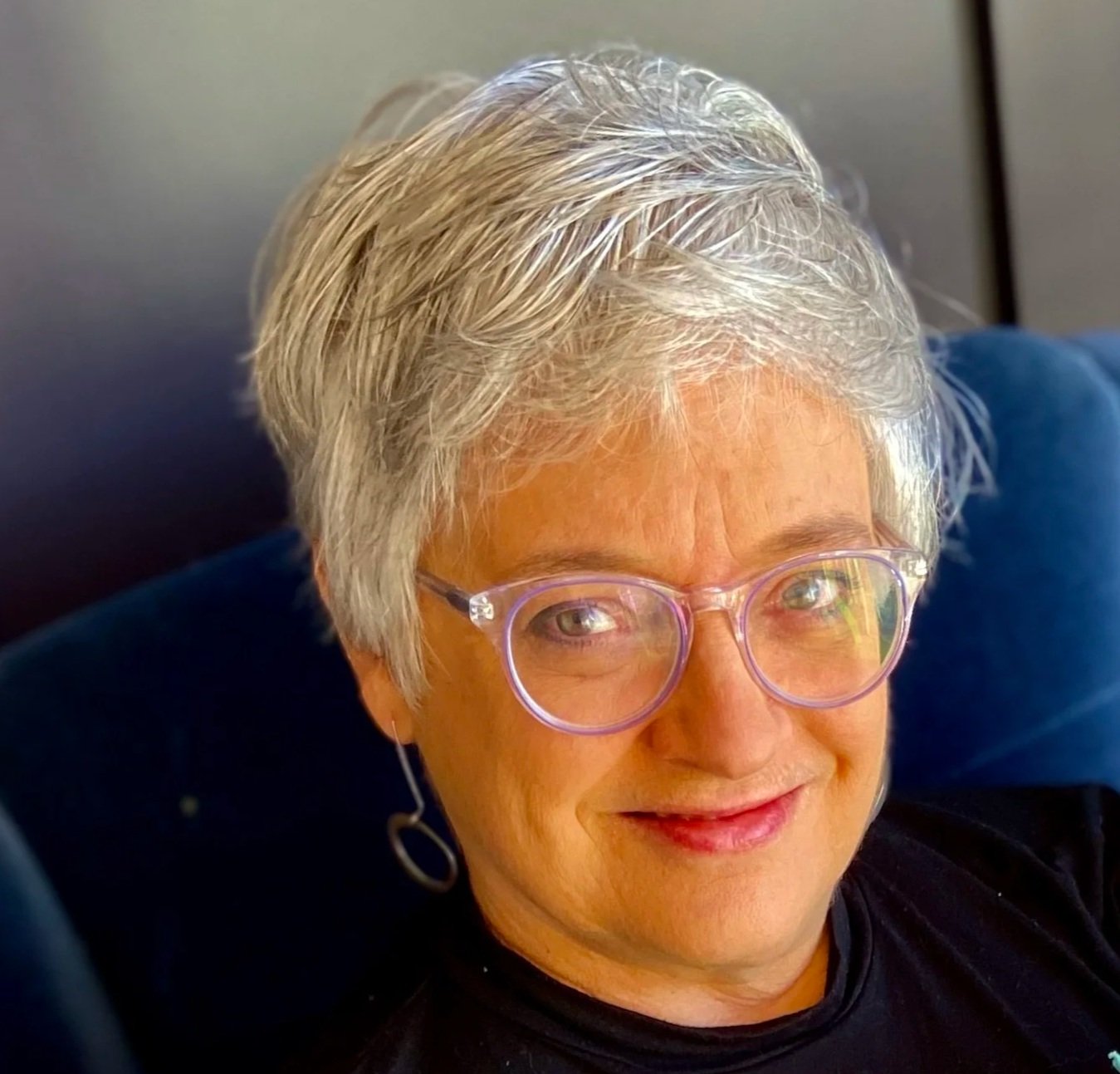Close-up of an older woman with short gray hair, pink glasses, and hoop earrings, smiling while looking at the camera.