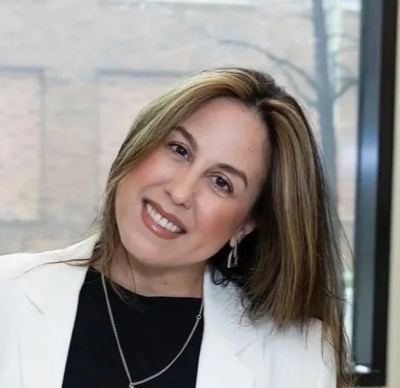 Woman with light brown hair, wearing a white blazer and black top, smiling and winking, standing indoors near a window.