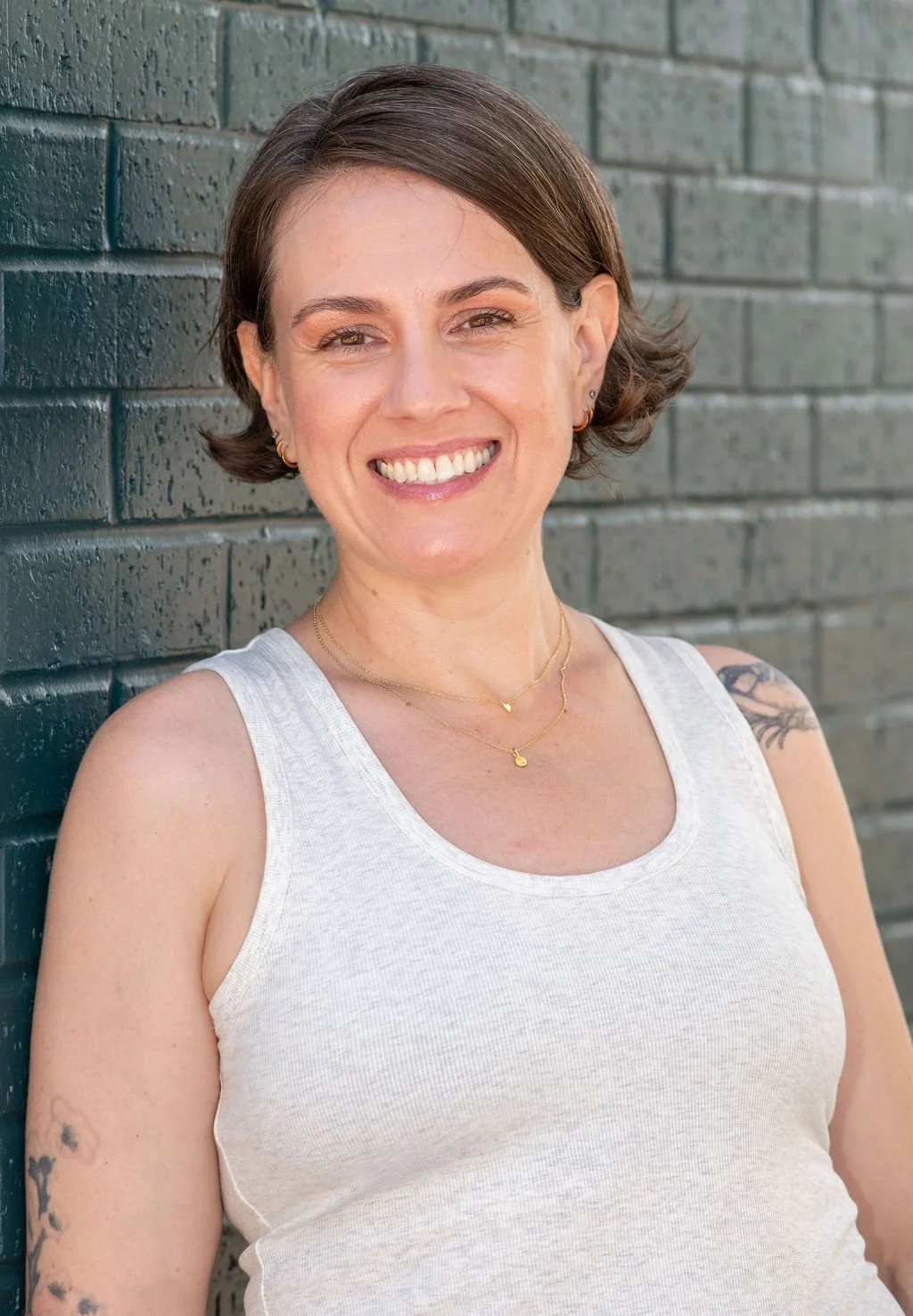 A smiling woman with short brown hair, wearing a white sleeveless top, gold jewelry, and tattoos on her arms, standing against a dark green brick wall.