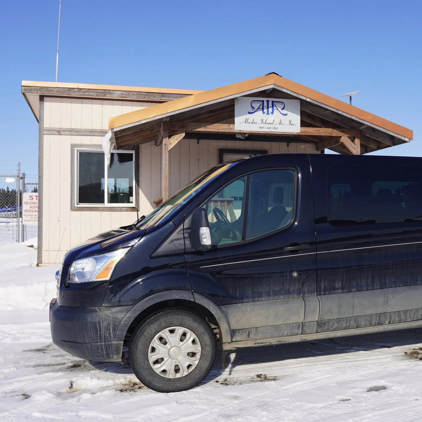 Black van parked in front of a building with a sign that reads 'Alaska Island Air, Inc.' The building has a small entrance with a wooden roof, and a window on the front wall. Snow covers the ground under the vehicle and around the building.