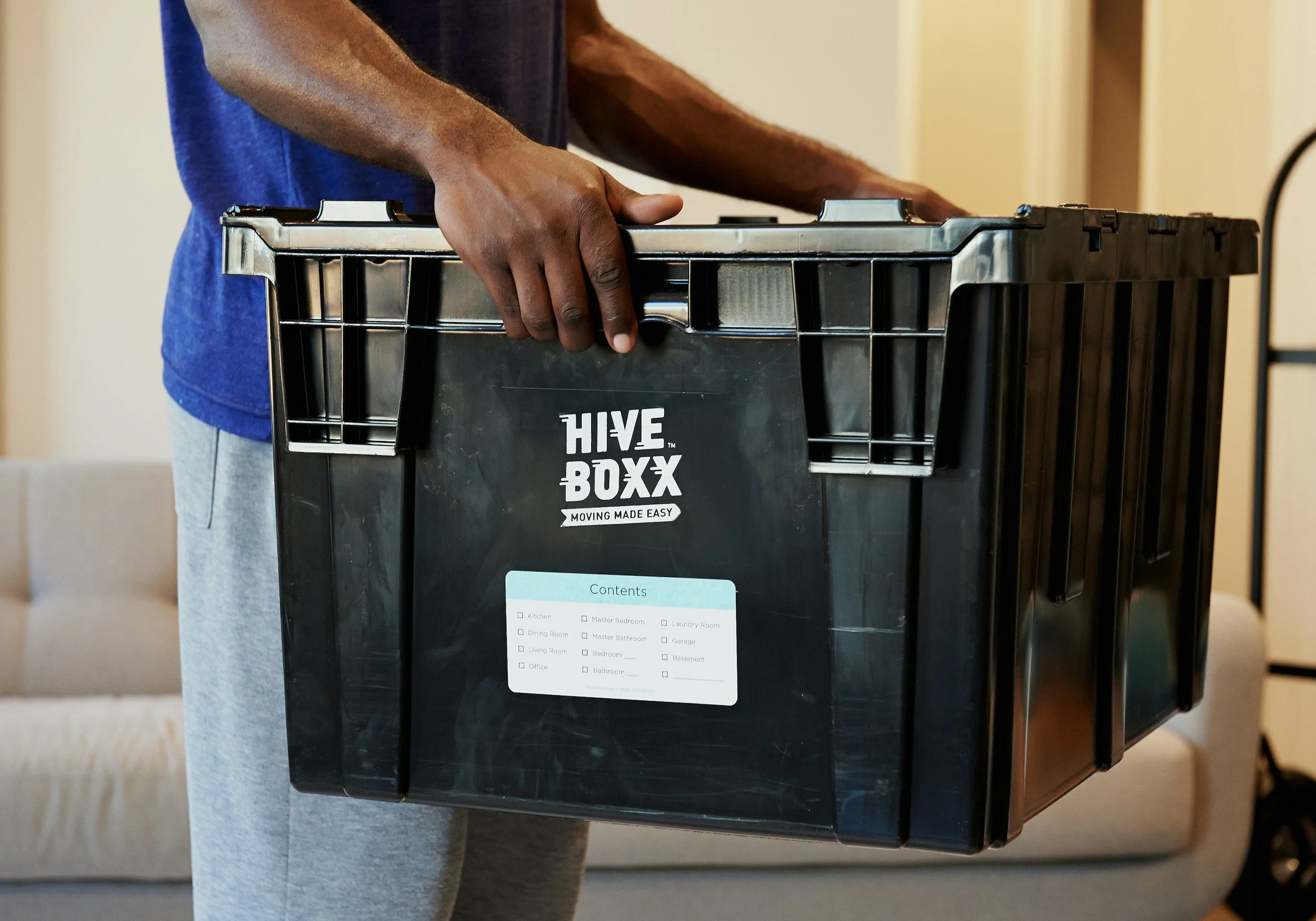 Person holding a black Hive box moving container with hand grip, standing indoors on a beige carpeted floor, partially shows a beige sofa in the background.