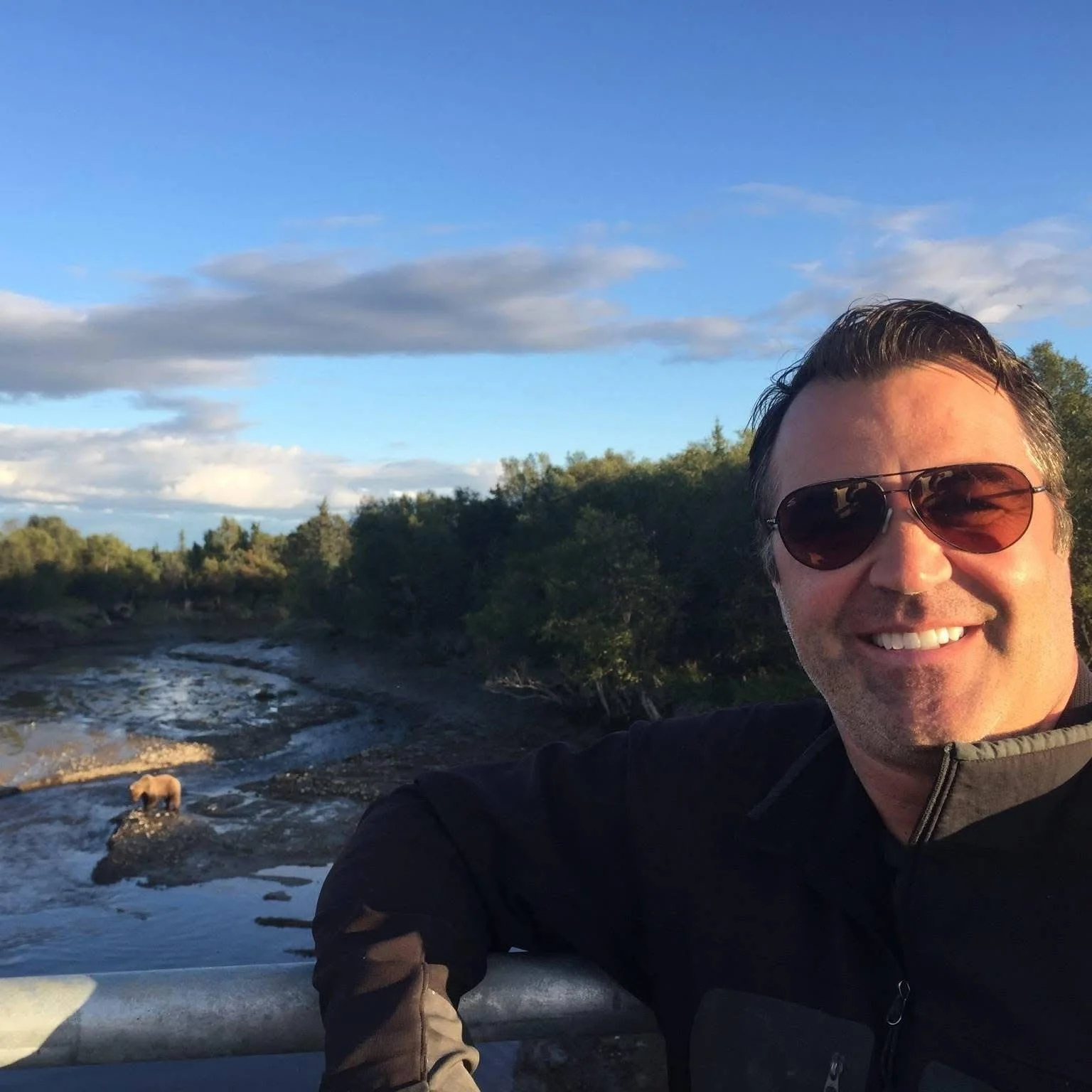 Smiling man wearing sunglasses taking a selfie outdoors with a river, trees, and a partly cloudy sky in the background.