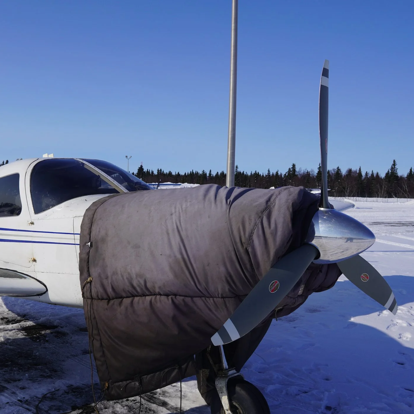Small airplane with a cover over the nose parked on a snow-covered tarmac, with a clear blue sky and a line of trees in the background.
