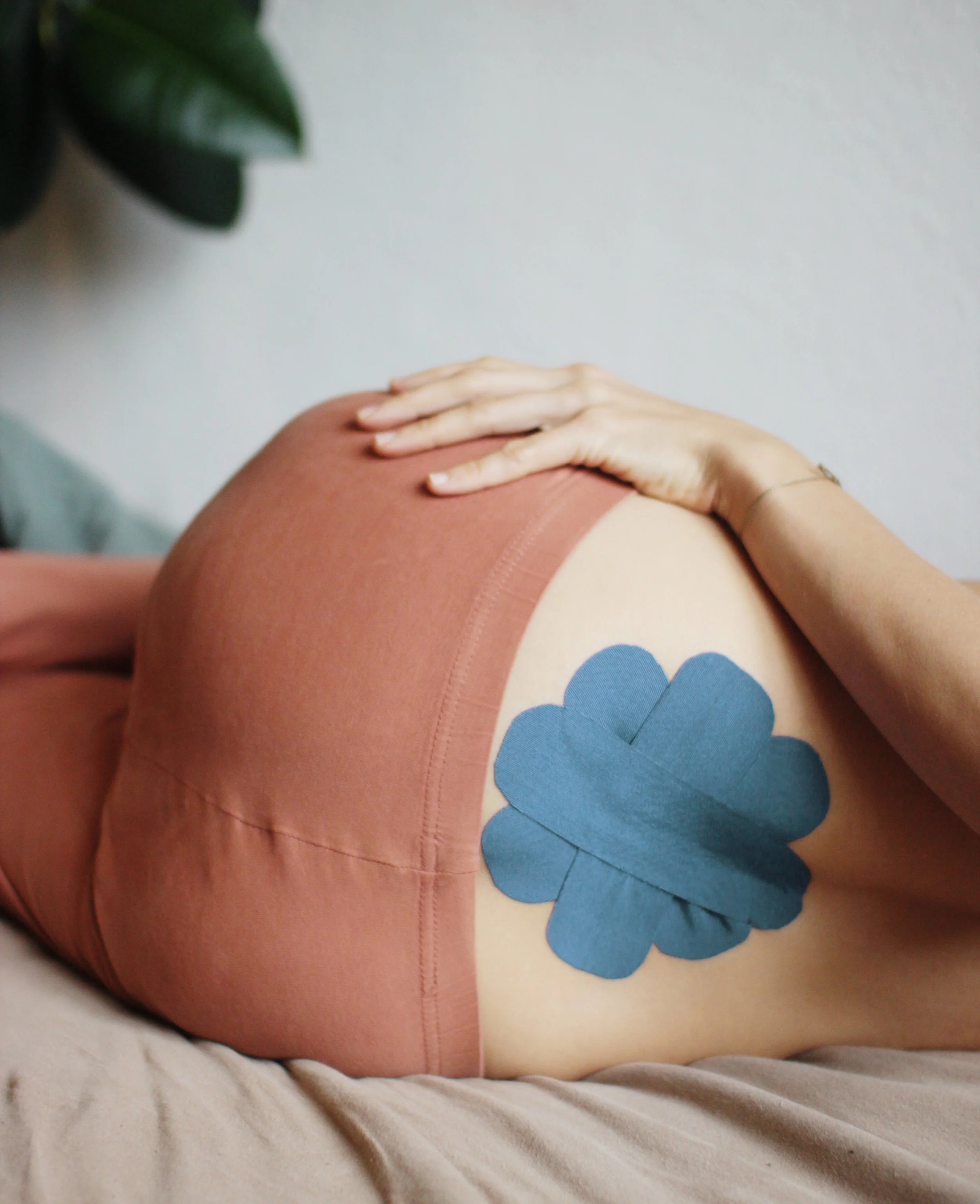 Rückseite einer schwangeren Frau, die auf einem Bett liegt. Blaues K-Tape (K-Taping) ist auf ihrem unteren Rücken in Blumenform befestigt.