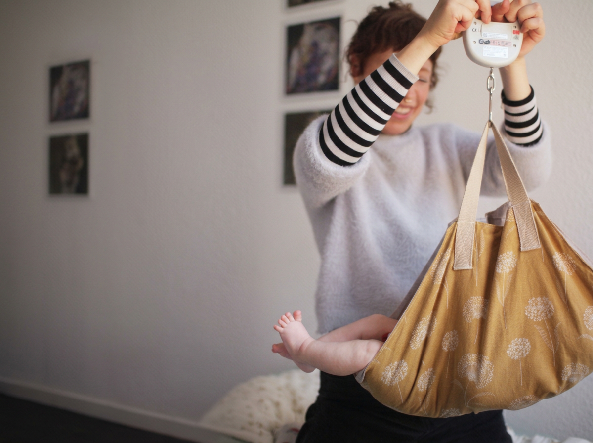 Hebamme Marie-Henrike Kruse hält eine Babywaage und ein gelbes Wiegetuch mit einem Baby darin. Aus dem Wiegetuch schauen die nackten Beine des Säuglings raus.