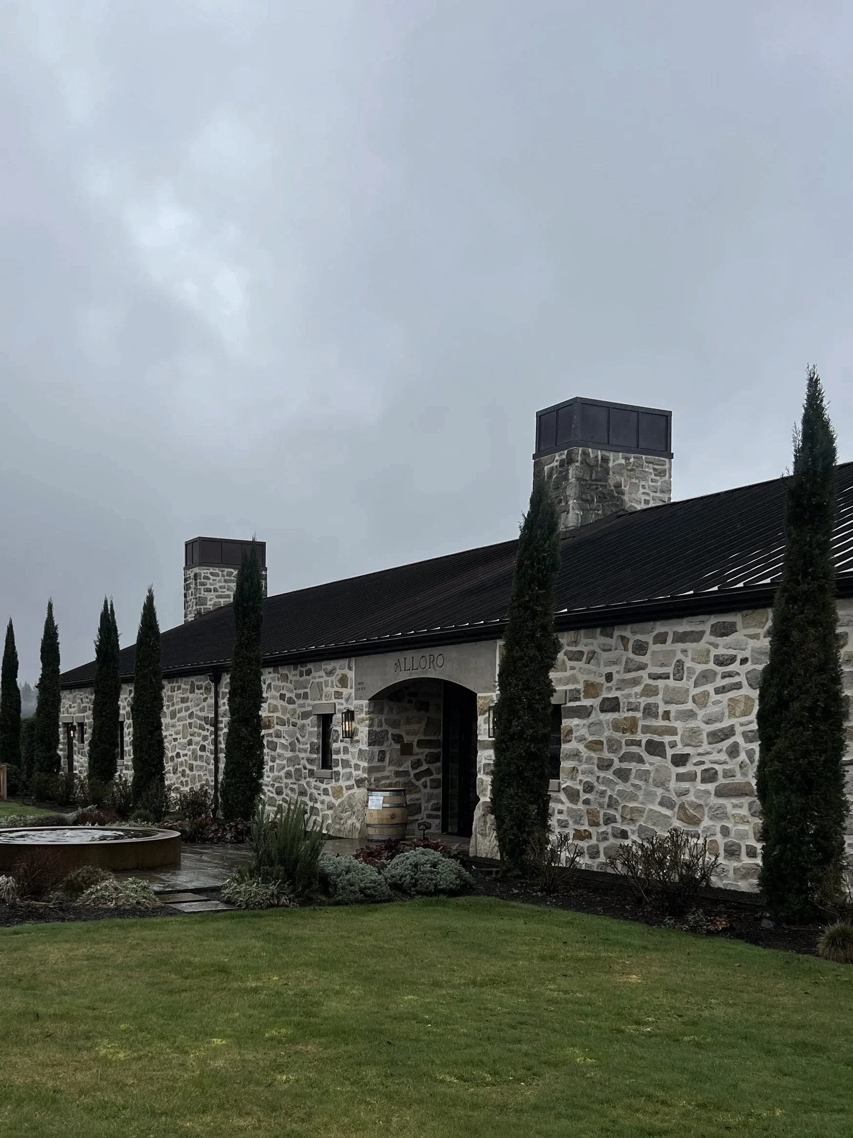A long and modern old world style stone building with cypress trees lining the front and arched entrance leading into Alloro Vineyard, Sherwood, Oregon