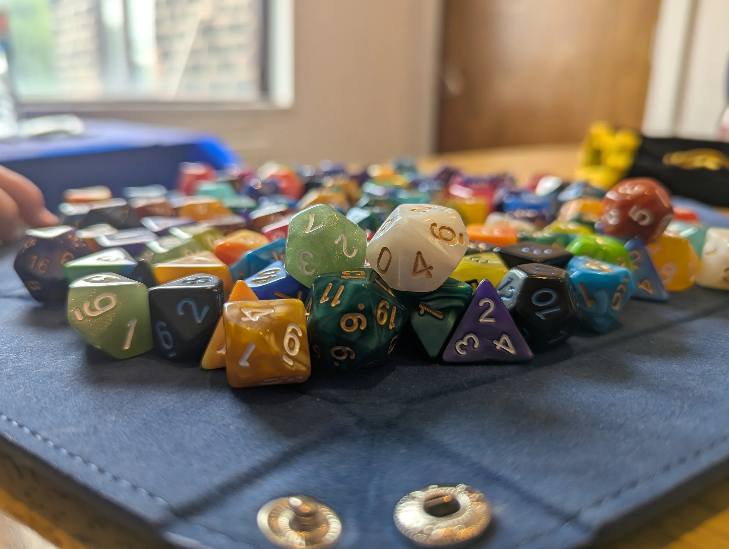 Assorted colorful polyhedral dice scattered on a black surface with a window in the background.