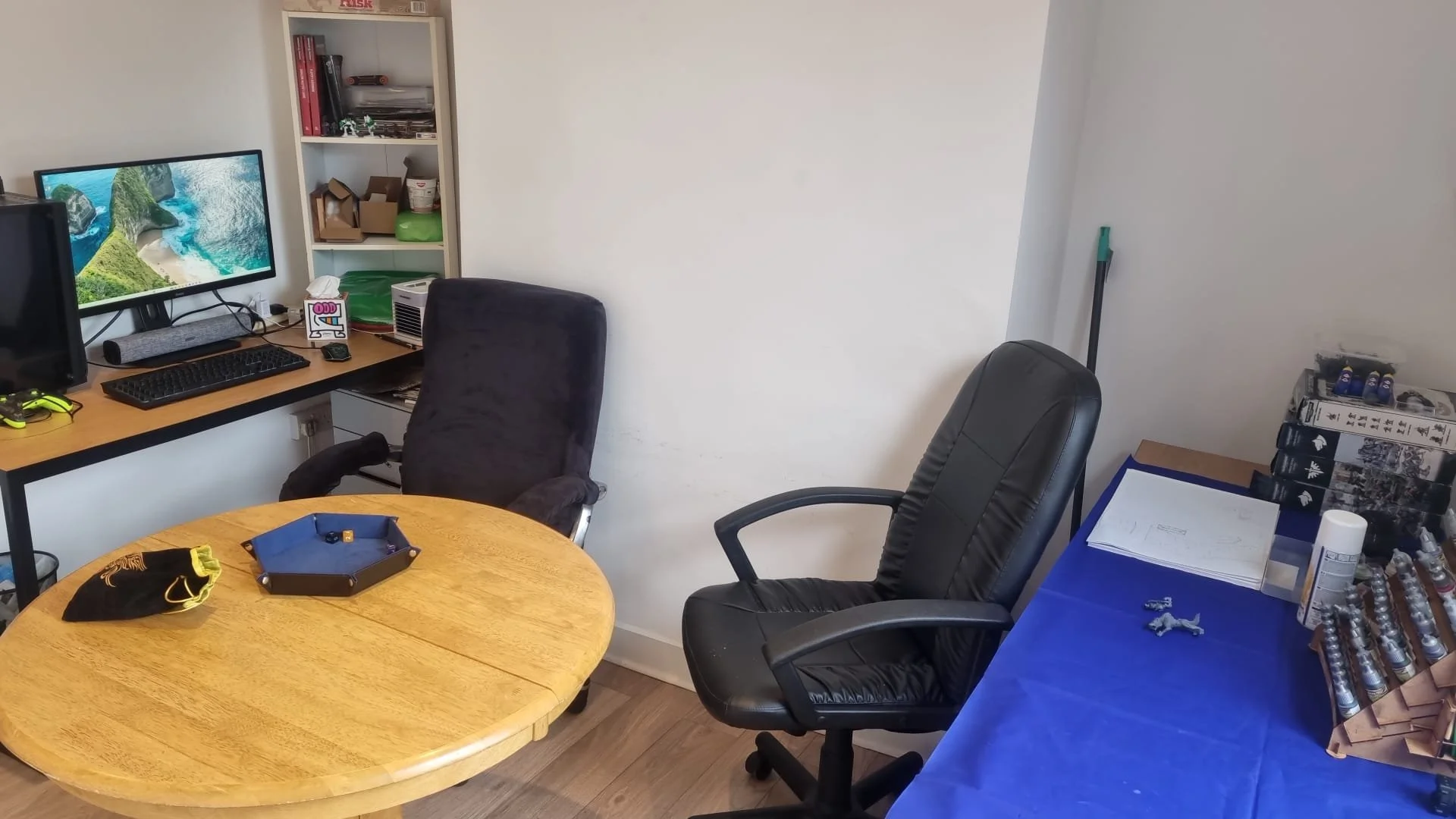 Office room with a round wooden table, two office chairs, a computer monitor showing a landscape, a bookshelf, and various office supplies.