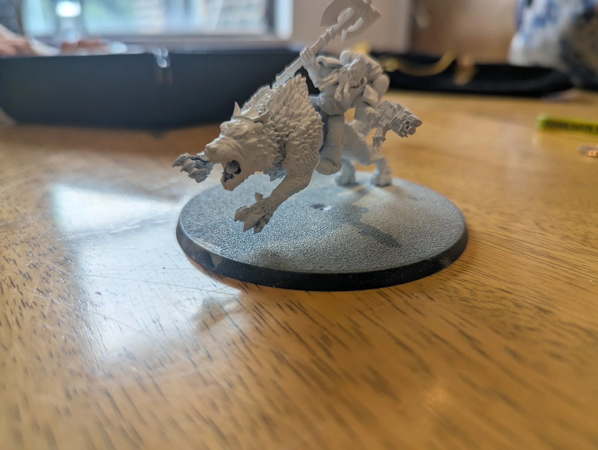 Unpainted miniature figurine of a dinosaur riding on the back of a warrior holding an axe, mounted on a round base, on a wooden surface.