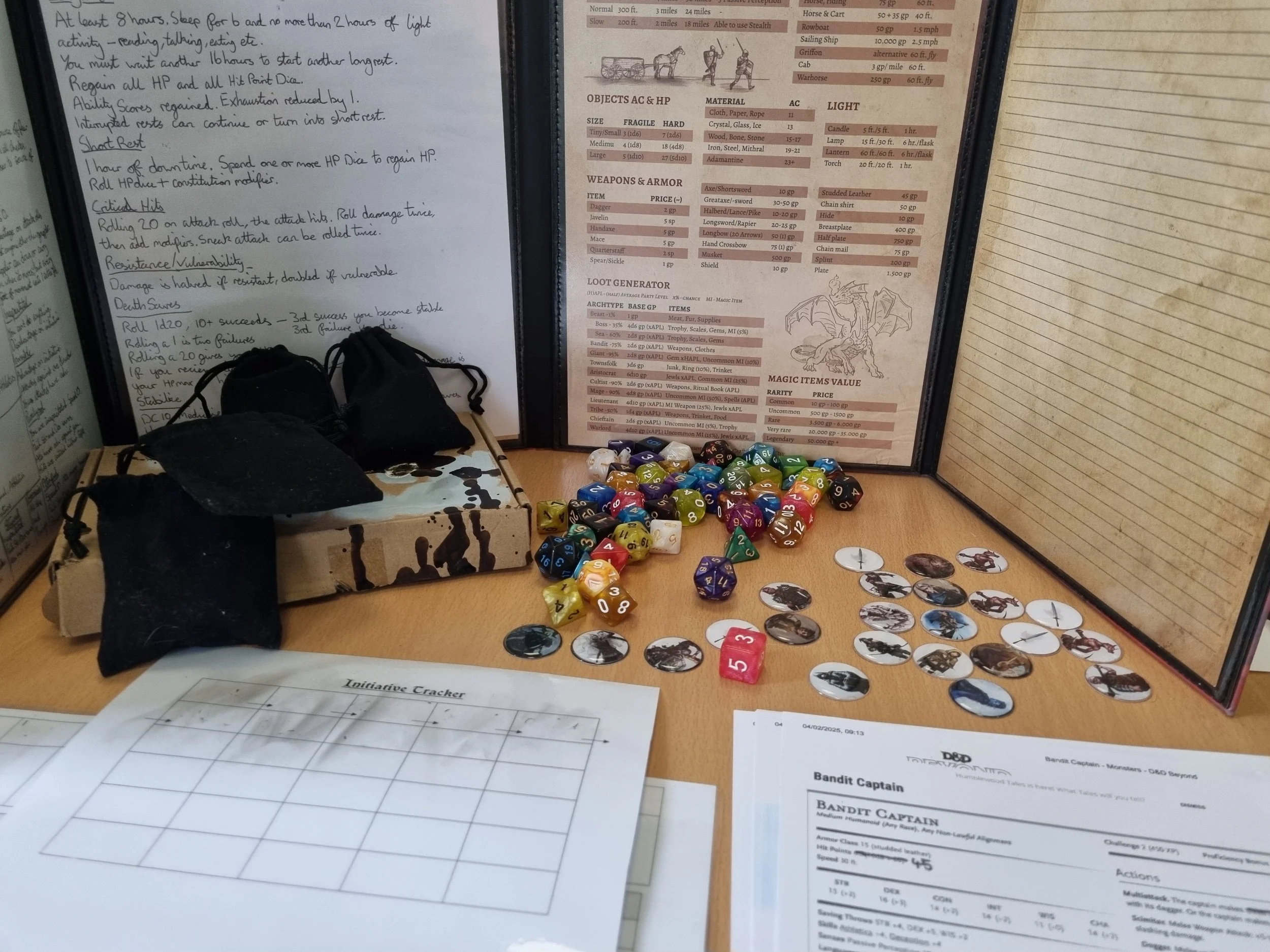 A tabletop role-playing game setup with colorful dice, character tokens, printed character sheets, and a handwritten notes sheet. There is also a cardboard box with black cloth bags. A detailed reference chart and a page with a dragon illustration are visible in the background.