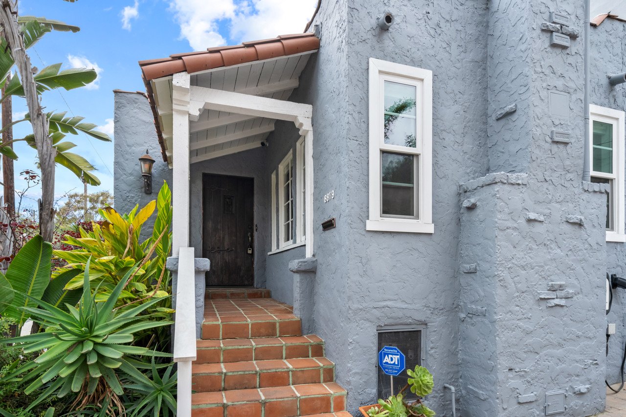 Exterior view of the a blue stucco bungalow with red brick tiles, a terracotta roof, and white trim.