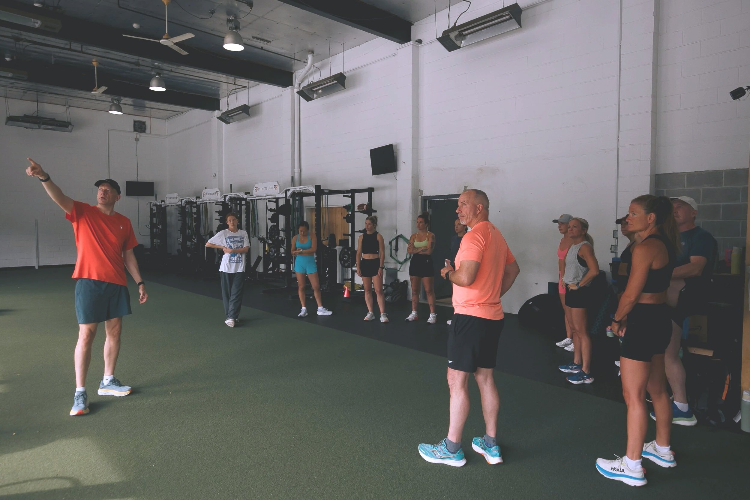 A fitness instructor leading a class of women in an indoor gym with white brick walls and gym equipment in the background.