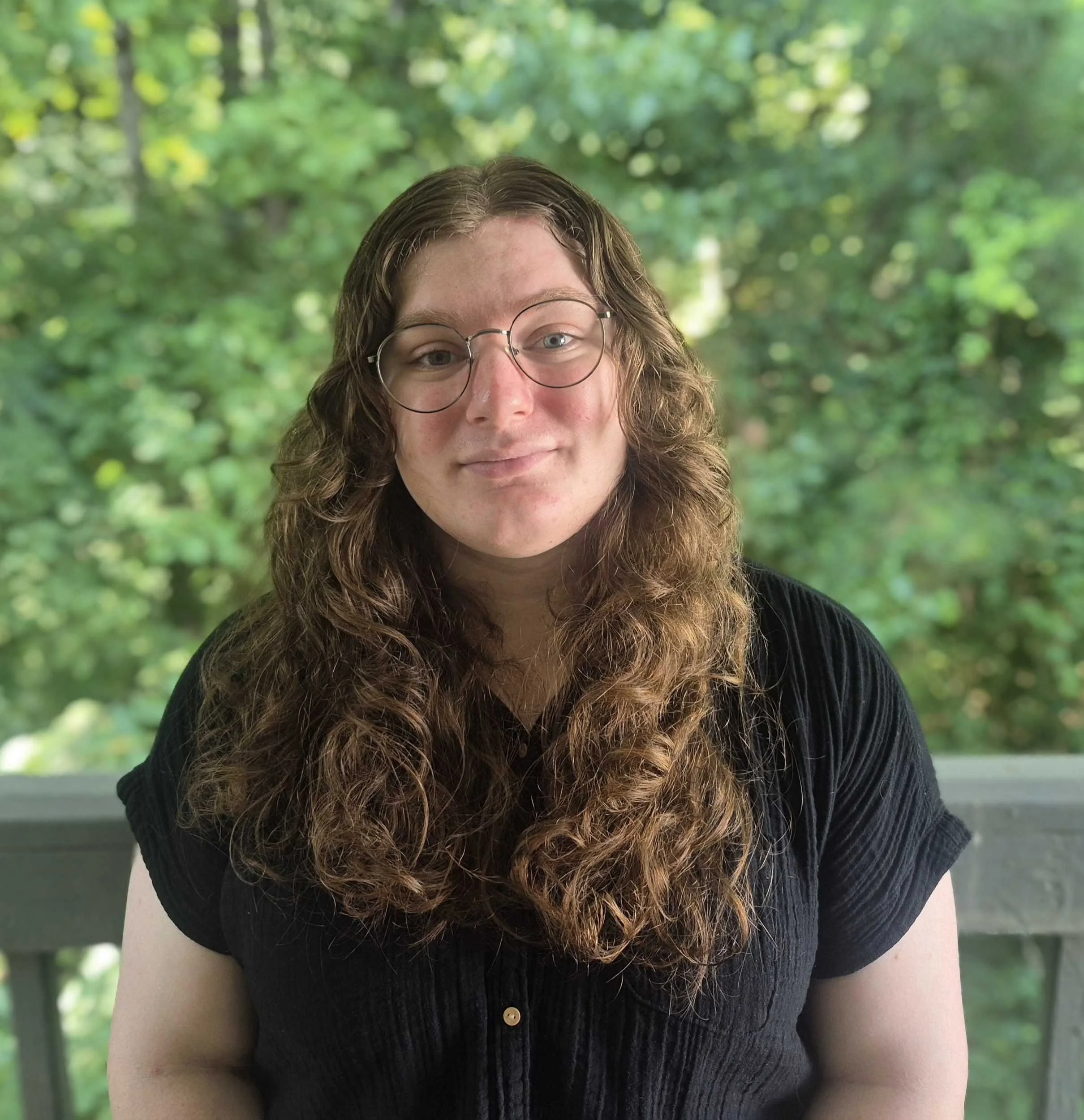 Bree presents a calm and cozy demeanor from a rustic park bench in a sunny wooded area. Her long hair rests elegantly over her shoulders. She is young, but flawlessly incorporates a homey 'cottage grandmother' vibe into her professional attire