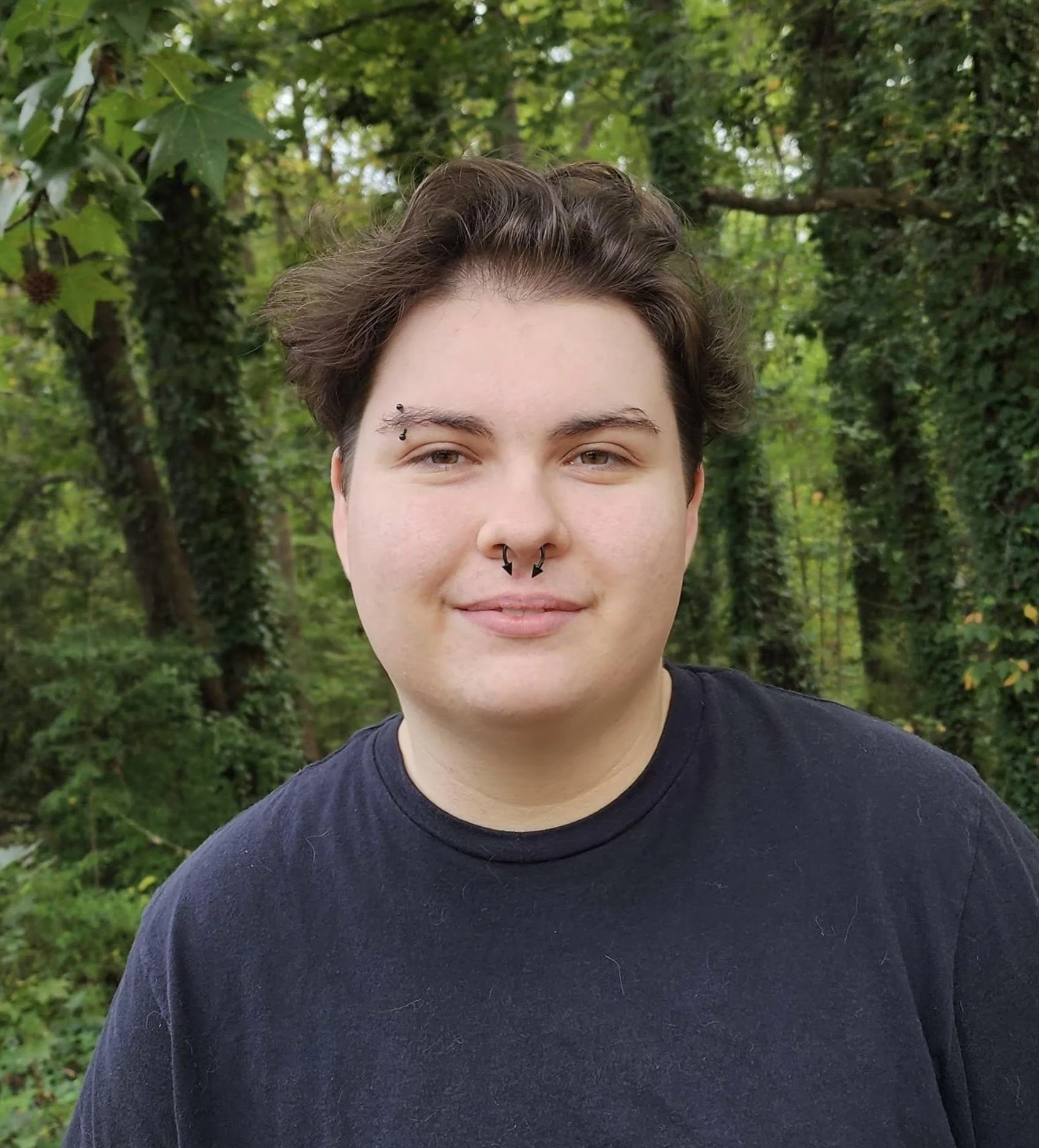 Cameron smiles reassuringly from a tranquil forest. His professional headshot playfully incorporates his piercings and swept back hair, while still staying grounded with his tasteful storm-grey color scheme