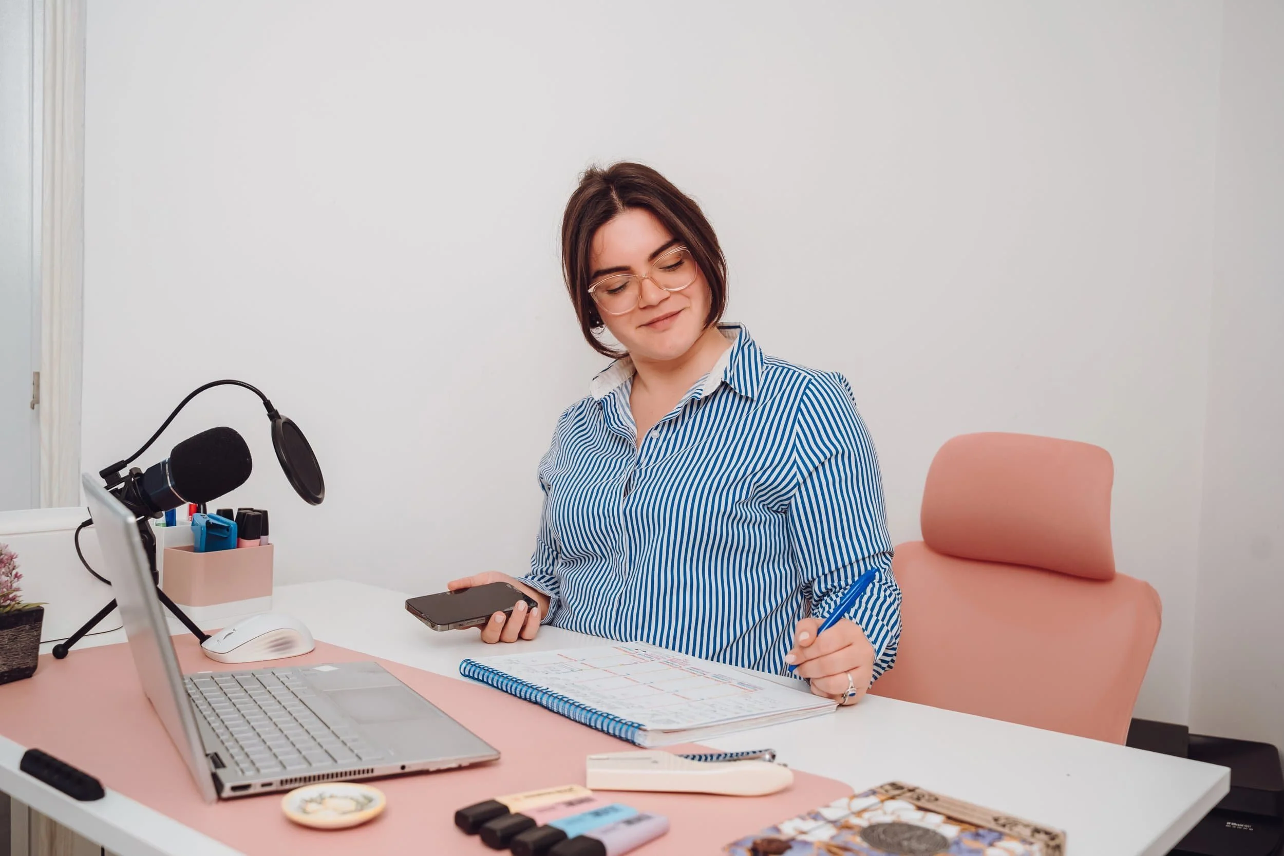 A young woman with short brown hair, glasses, and a striped blue and white shirt sits at a desk, looking at her phone with a slight smile, surrounded by office supplies and a laptop.