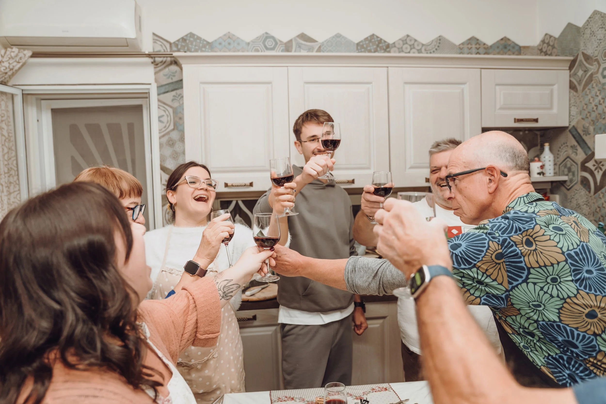 Two women sitting at an outdoor table, smiling and talking to each other, with plates, bowls, a teapot, and a notebook on the table, surrounded by flowering plants and trees.