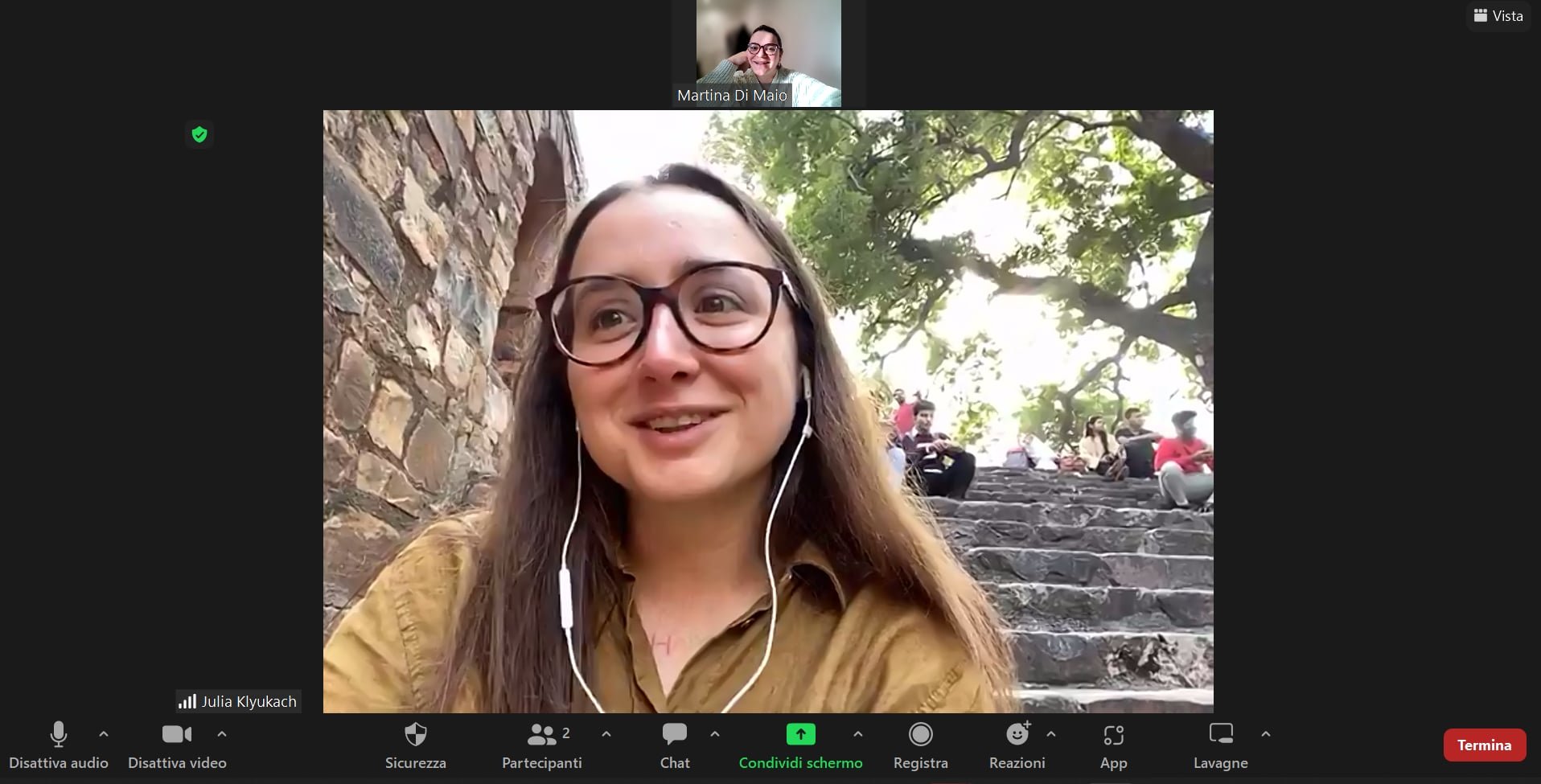 Zoom video call showing a woman with glasses and long hair smiling outdoors, with stone steps and large trees in the background. Other people sit on the steps.