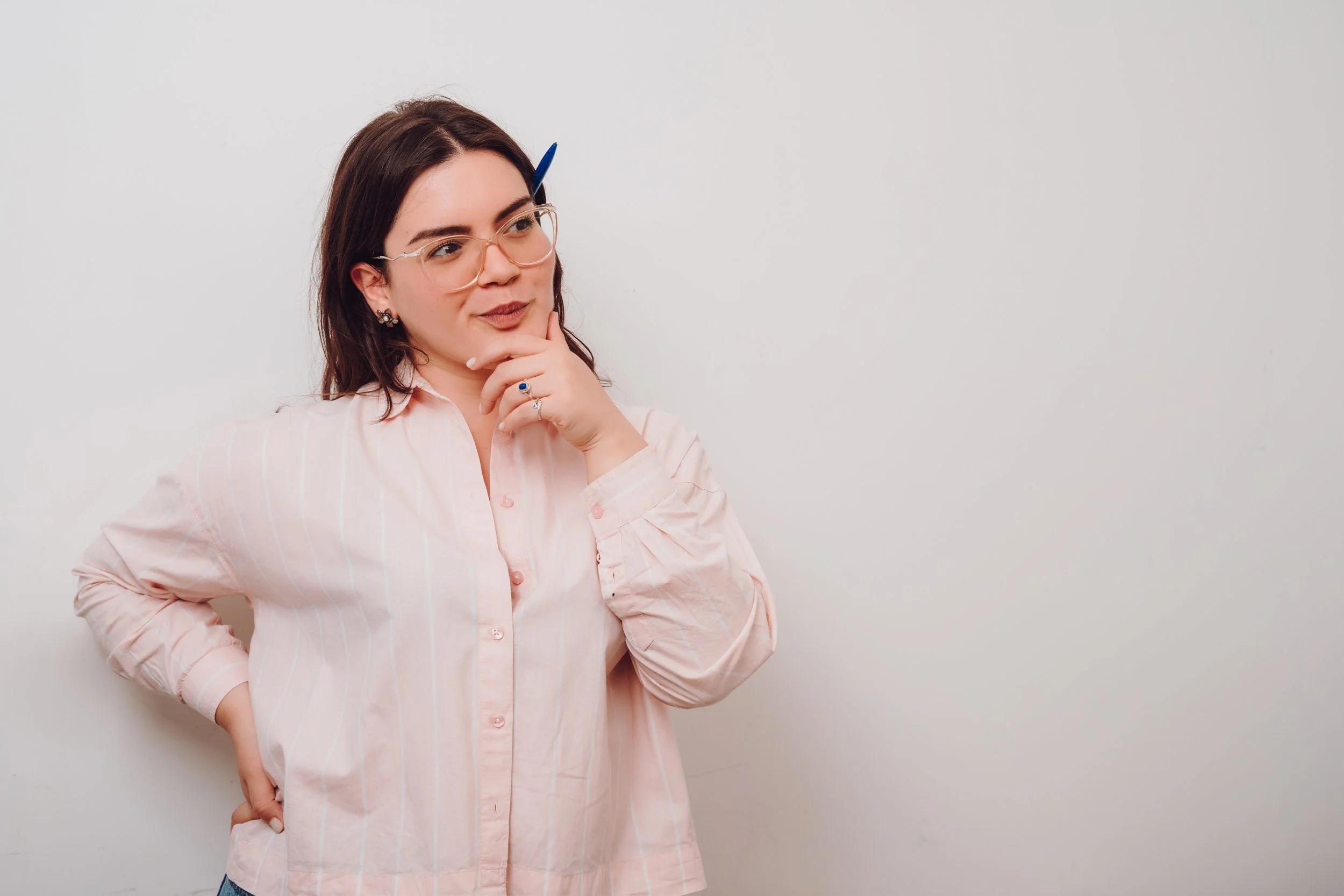 Woman with dark hair, glasses, and a pink blouse, standing against a white wall with a thoughtful expression.