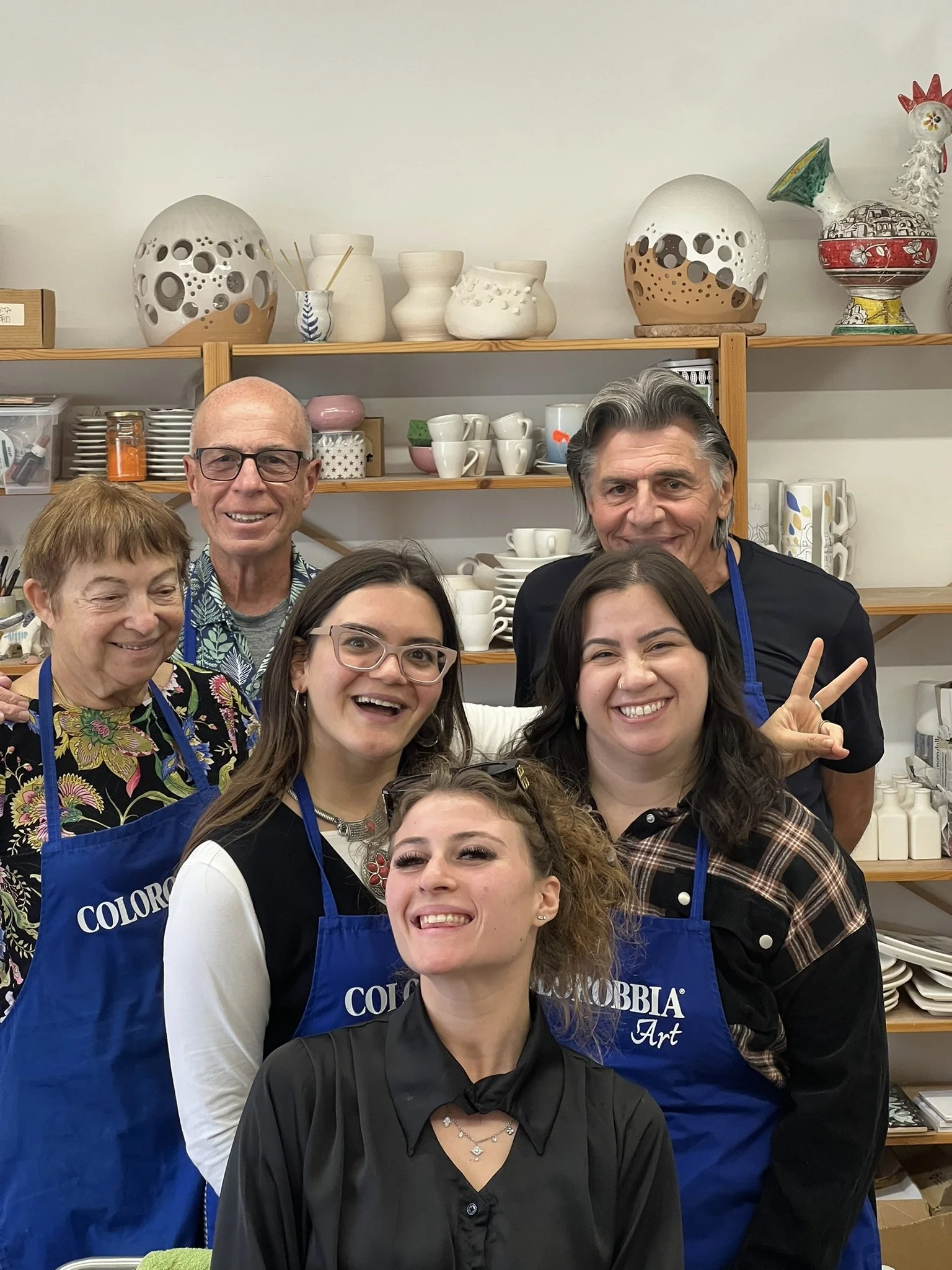 Two women sitting at a table, smiling and working together with papers, notebooks, a laptop, and coffee cups in a well-lit room with lace curtains.