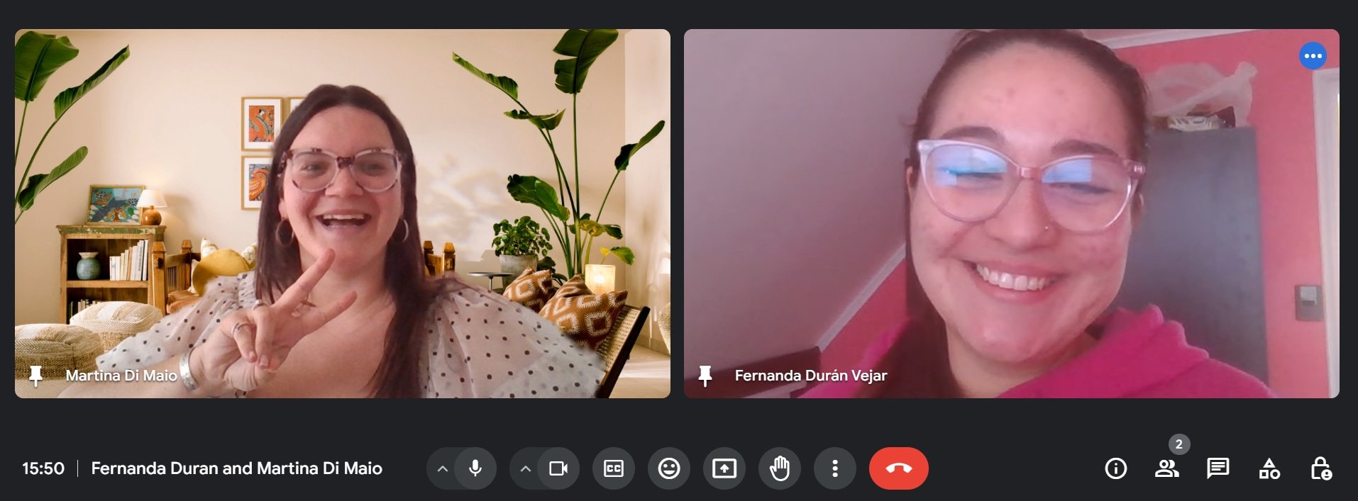 Two women smiling during a video call, one is wearing glasses and a pink hoodie, the other has glasses and is making a peace sign, with a warmly decorated room in the background.