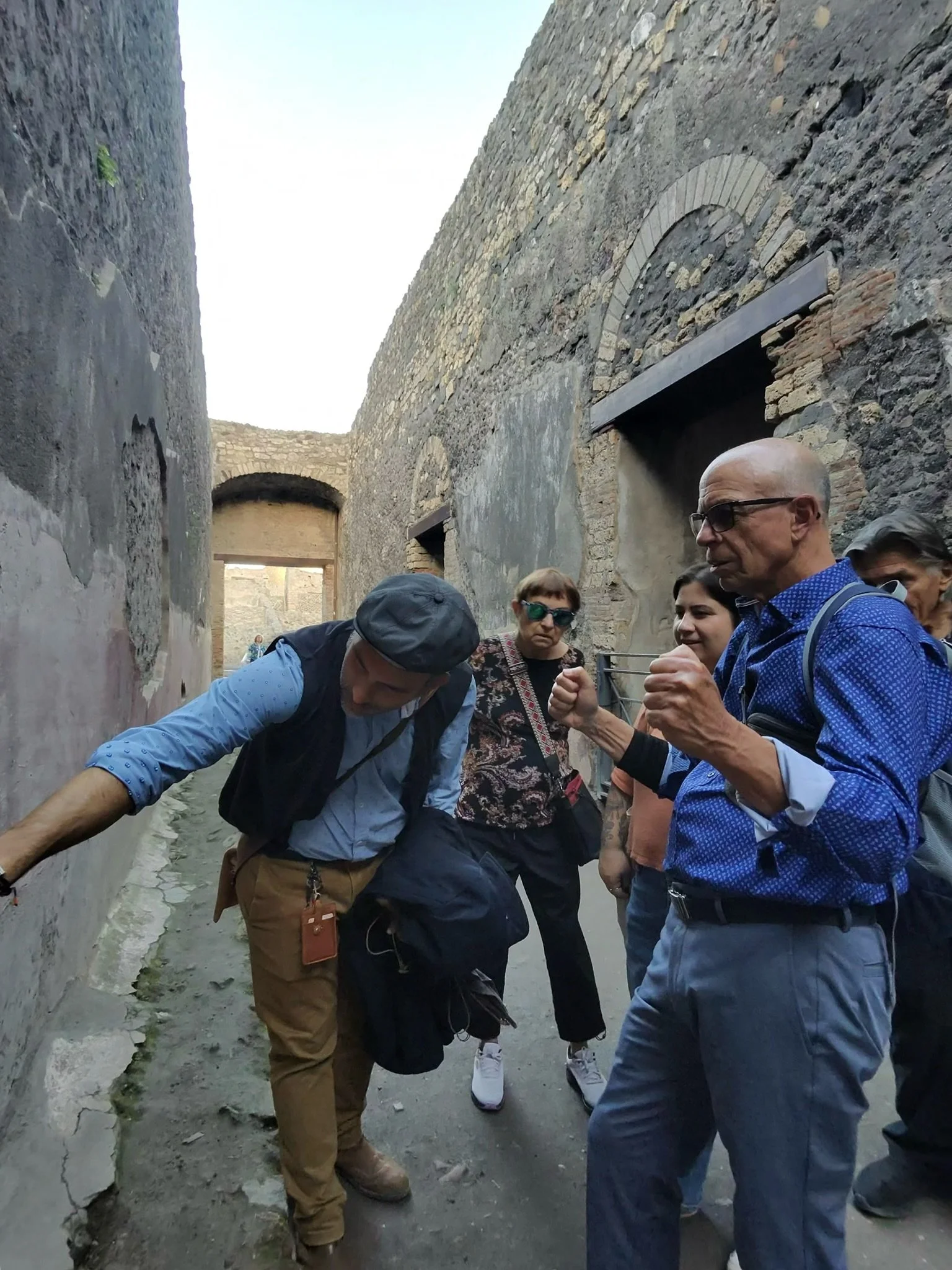 Two women sitting at an outdoor table, smiling and talking to each other, with plates, bowls, a teapot, and a notebook on the table, surrounded by flowering plants and trees.