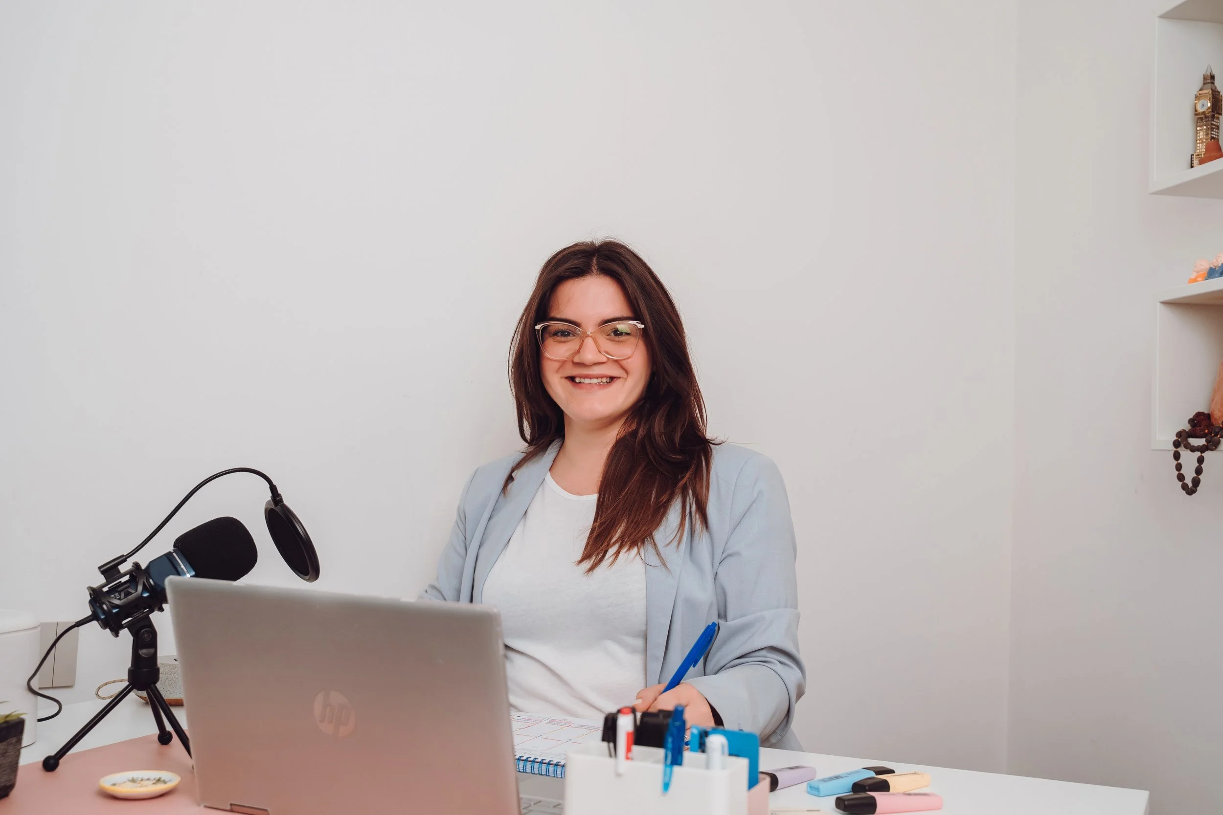 A woman with glasses sitting at a desk with a laptop, microphone, and stationery, smiling at the camera.