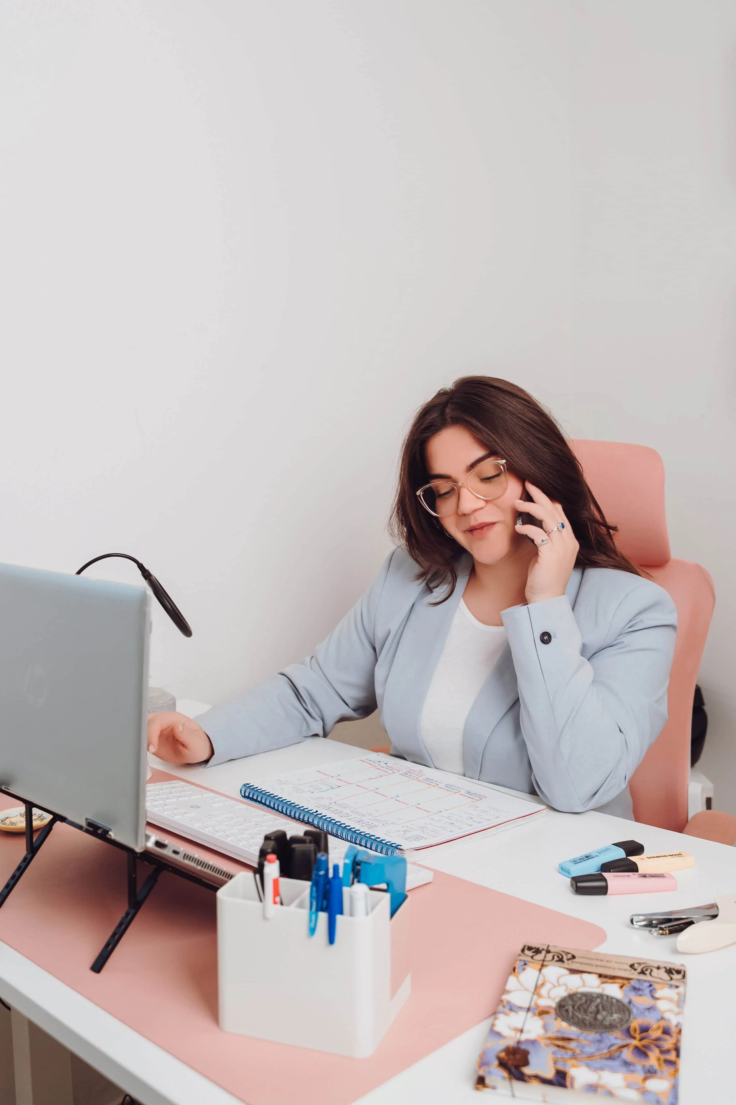 A woman with glasses, wearing a light gray blazer, sitting at a white desk with pink accents, smiling and talking on the phone. The desk has various office supplies including markers, a notebook, a computer monitor, and a planner.