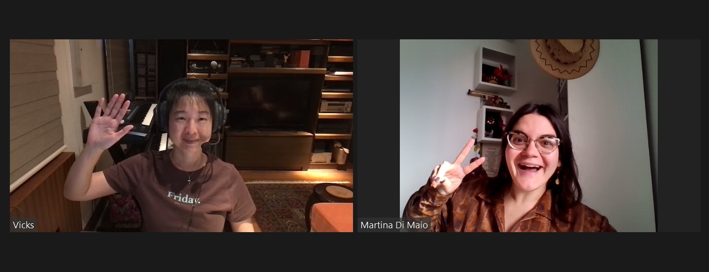 Two women on a video call, both smiling and making peace signs. The woman on the left has short dark hair, wearing headphones and a brown shirt, sitting in a room with a piano and shelves. The woman on the right has shoulder-length dark hair, glasses
