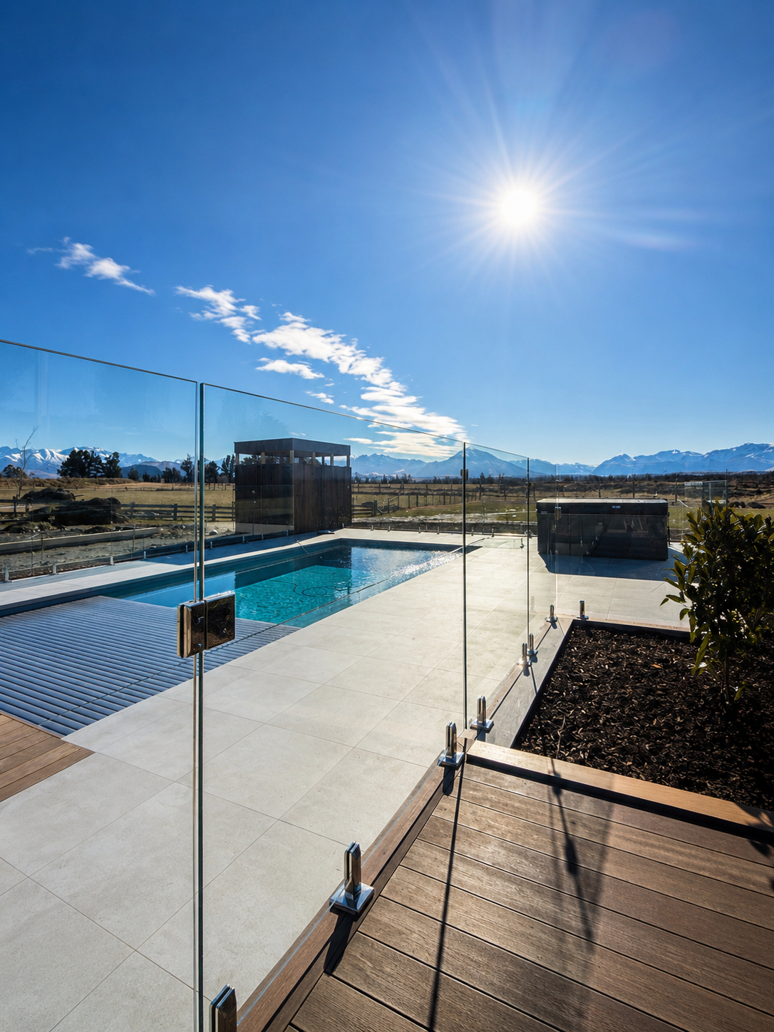 A backyard with a small rectangular swimming pool, enclosed by a glass fence, with mountains in the background under a bright sunny sky.