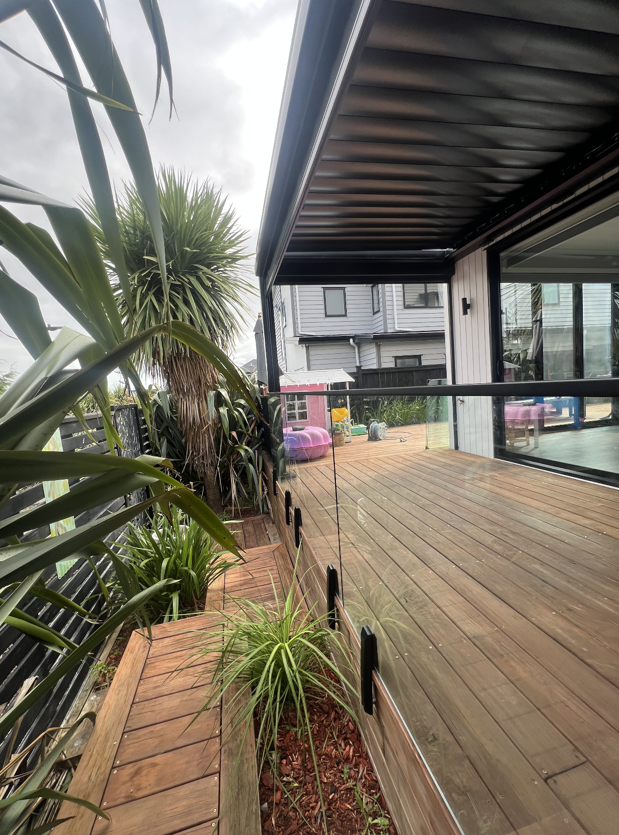 A wooden backyard deck with a clear glass safety barrier, surrounded by lush green plants, and a neighboring house in the background under a cloudy sky.