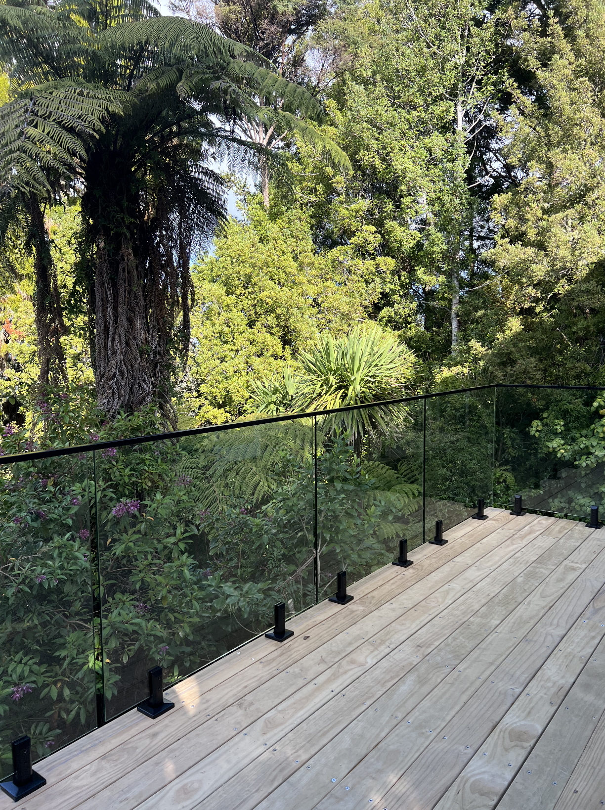 View of a wooden deck with glass railing, overlooking lush green trees and plants in a forest or garden setting.