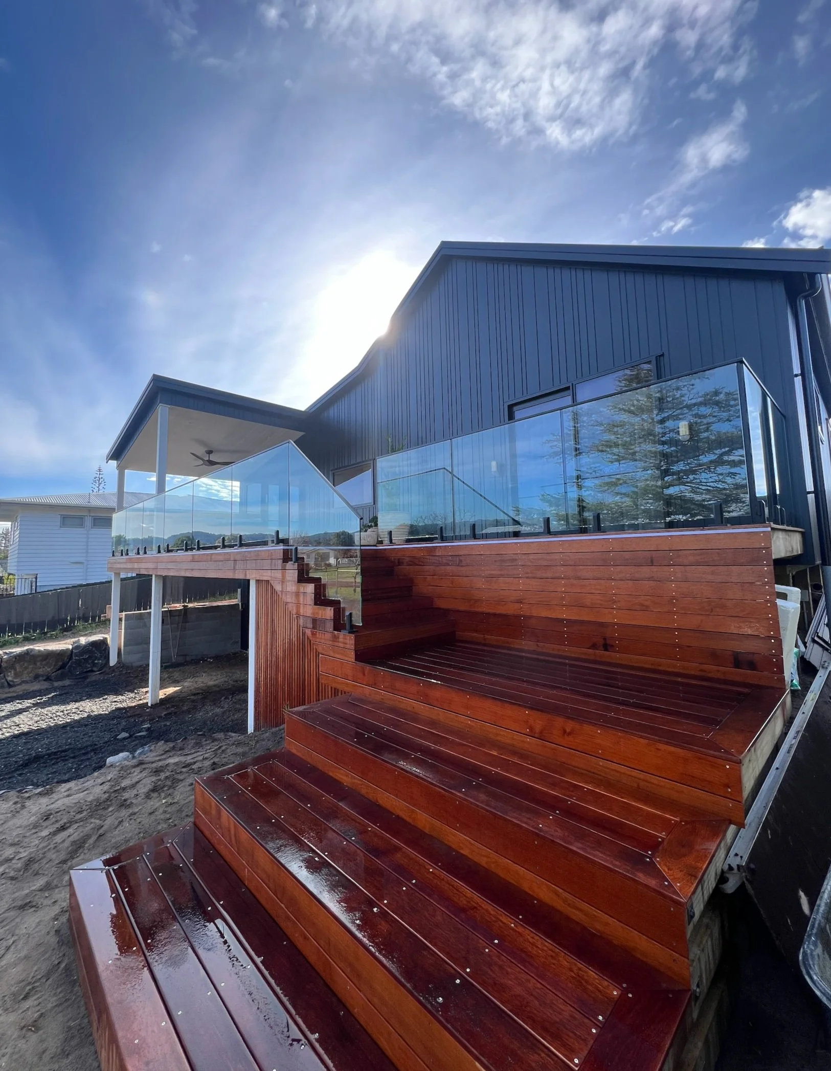 New wooden outdoor deck with glass railing attached to a modern blue house, under a partly cloudy sky.