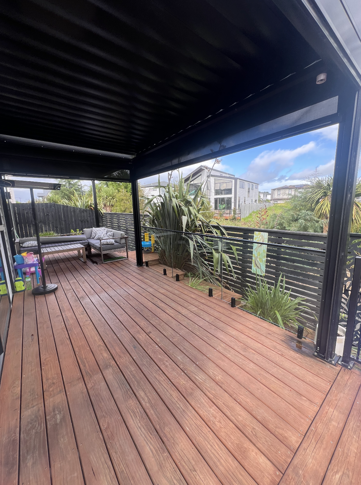 An outdoor wooden deck with modern railing, some outdoor furniture, and large plants, under a black metal roof, with neighboring houses and a partly cloudy sky in the background.