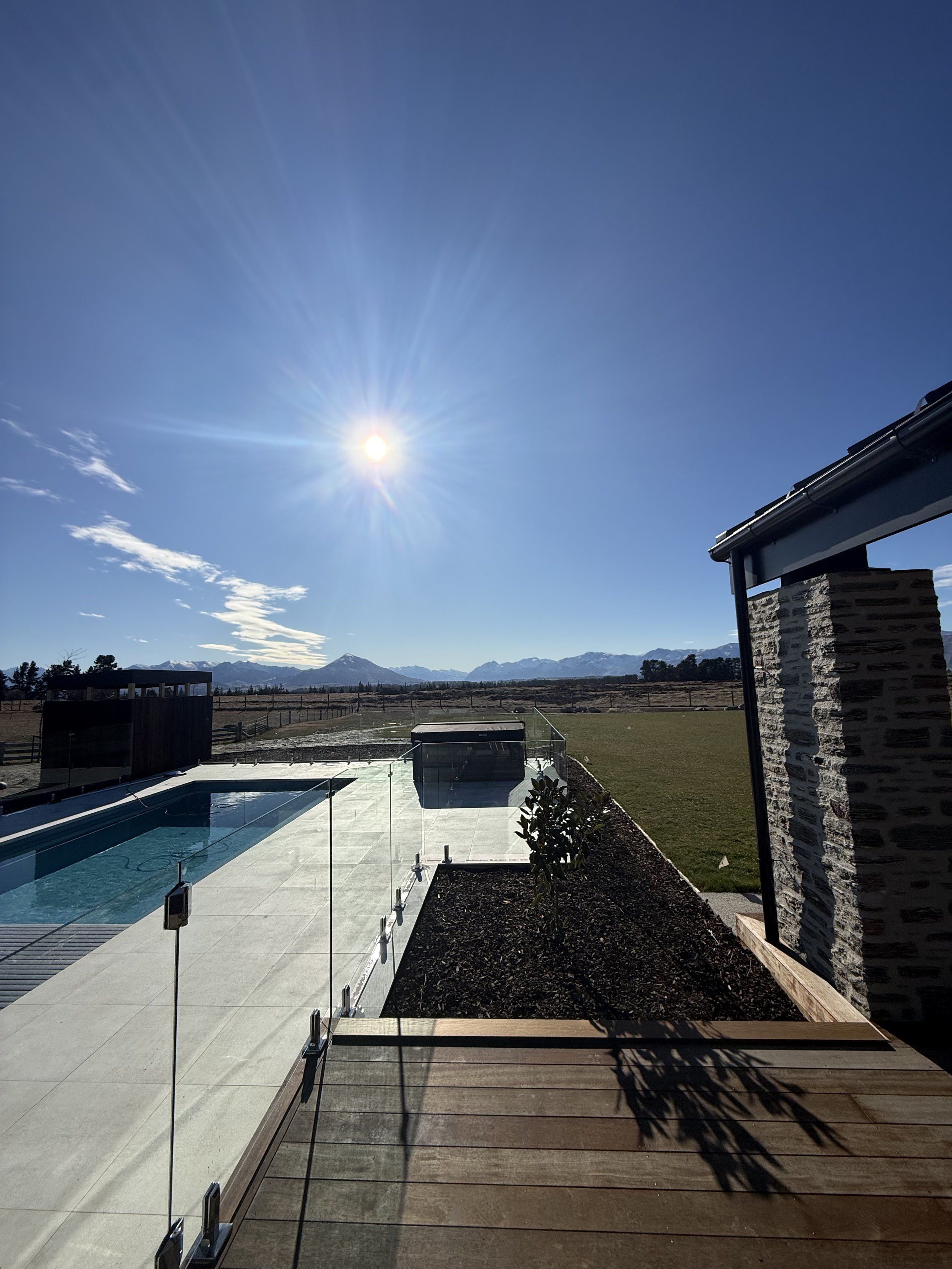 A backyard scene showing a swimming pool with a glass fence, a grassy area, and mountain range in the distance. The sky is clear with the sun shining brightly.