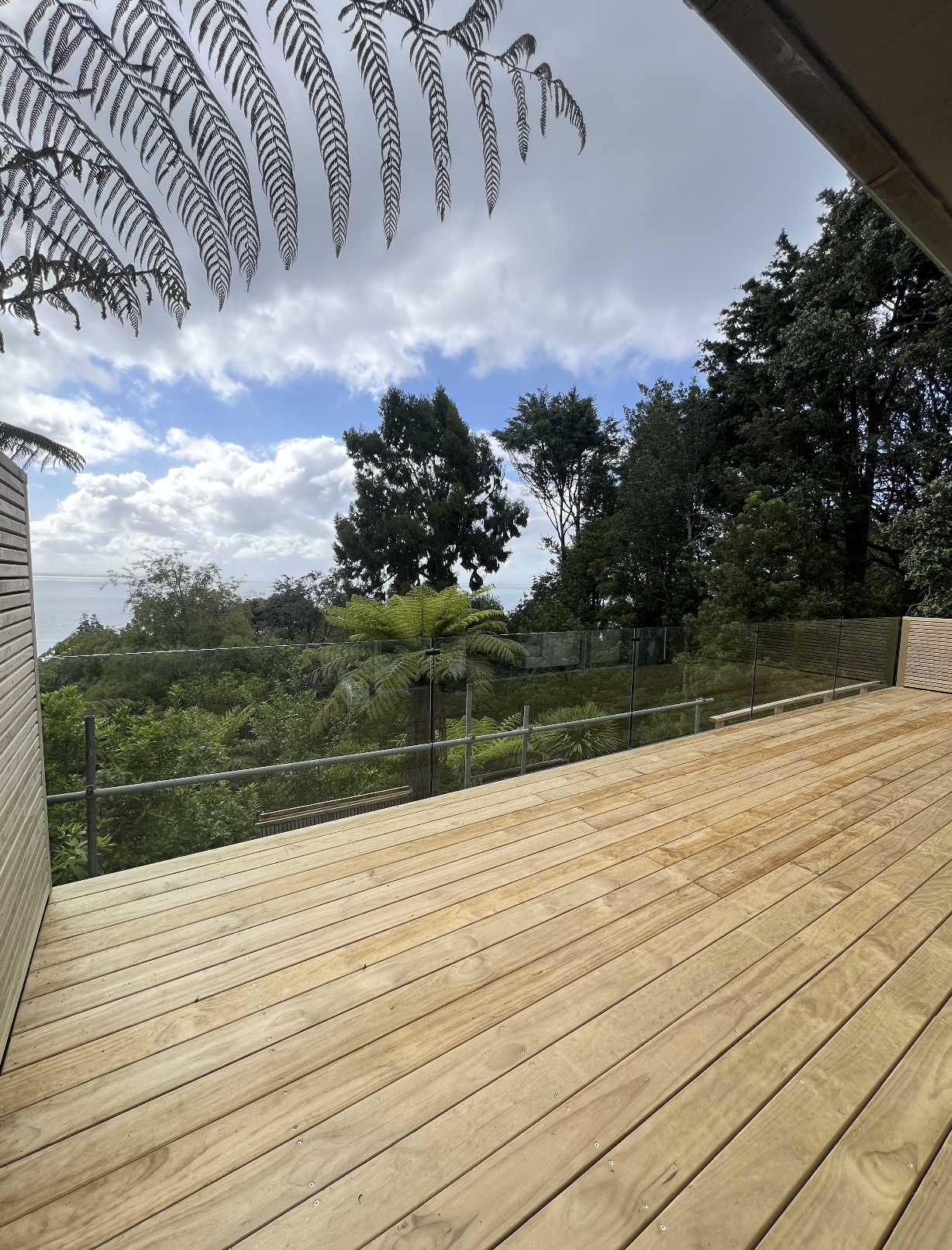 View of a wooden deck with glass railing overlooking lush green trees and a partly cloudy sky.