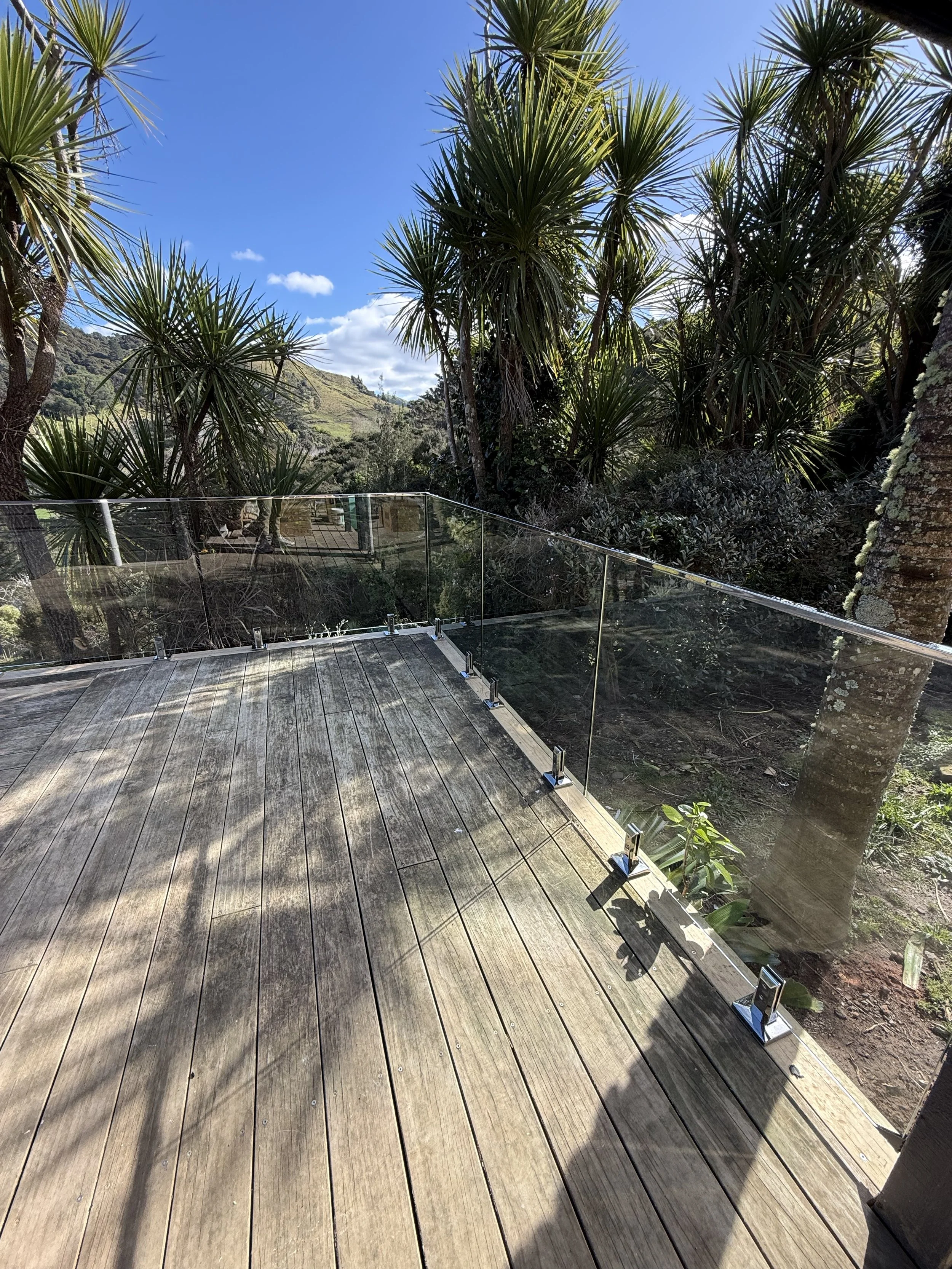 A wooden deck with glass railing overlooking lush green trees and a hill under a bright blue sky with a few clouds.
