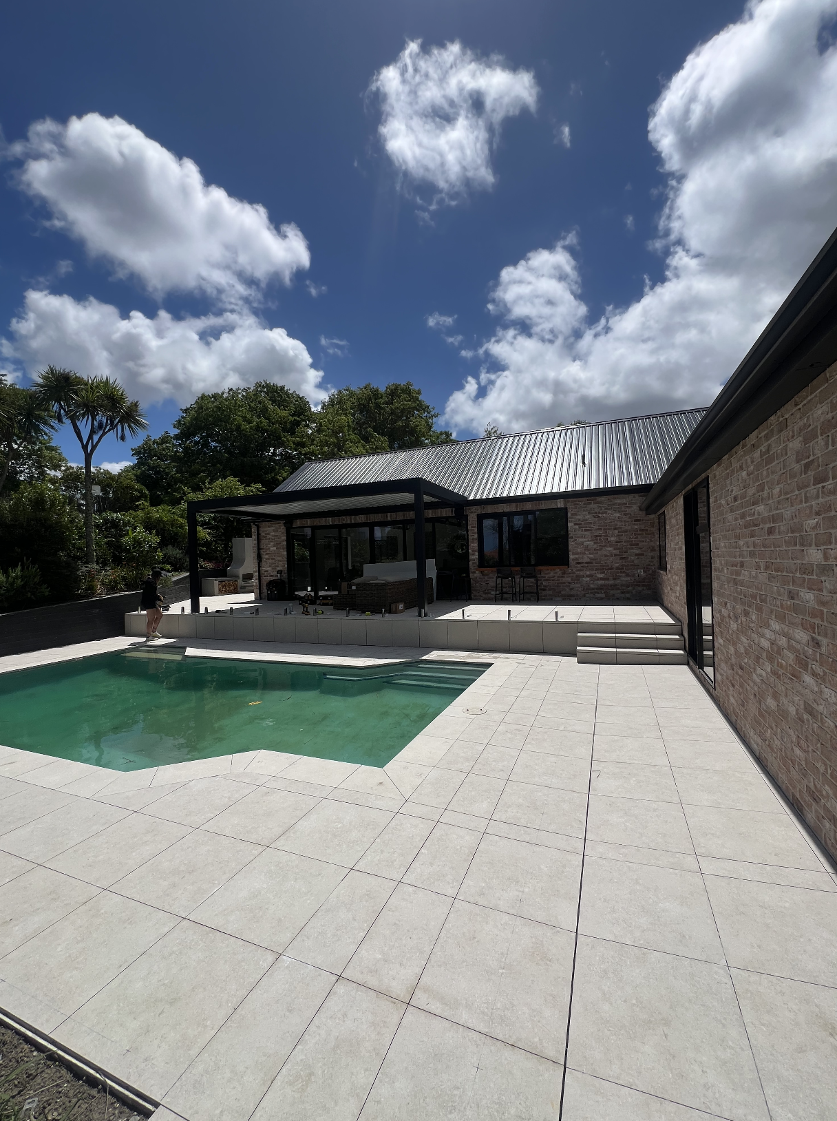 A backyard with a swimming pool, a house with brick walls, a covered patio area with outdoor furniture, and steps leading up to the outdoor space. Trees and a partly cloudy sky are visible in the background.