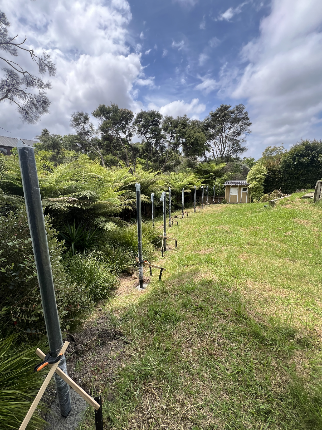 A backyard with a grass lawn and a row of metal poles with brackets, likely for a structure, on the left side. Green bushes and trees border the yard, and a small shed is visible in the distance. The sky is partly cloudy with patches of blue.