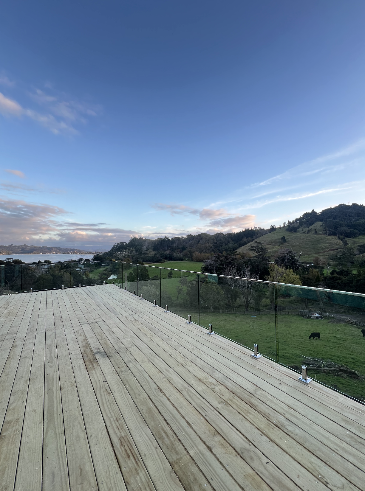 A view of a wooden deck with glass railing overlooking green hills, trees, water, and a blue sky with some clouds.