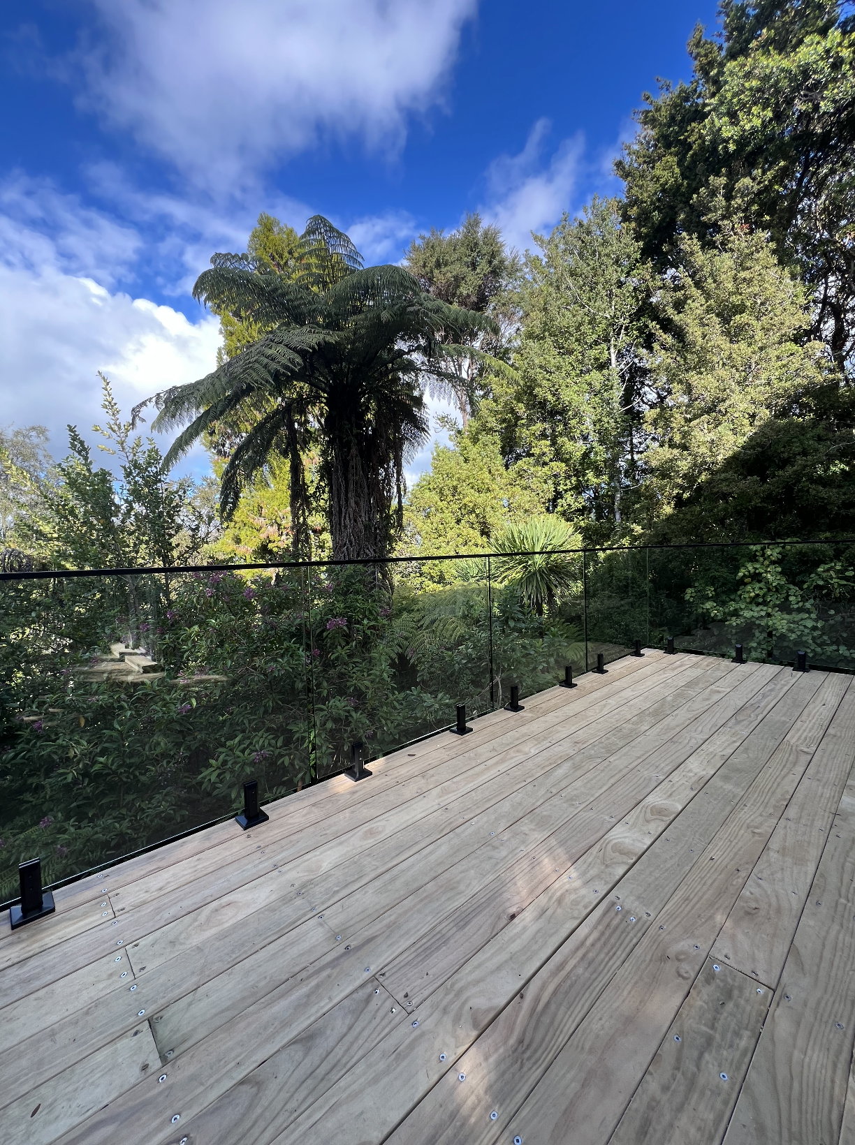 Wooden deck overlooking lush green trees and a bright blue sky with scattered clouds.
