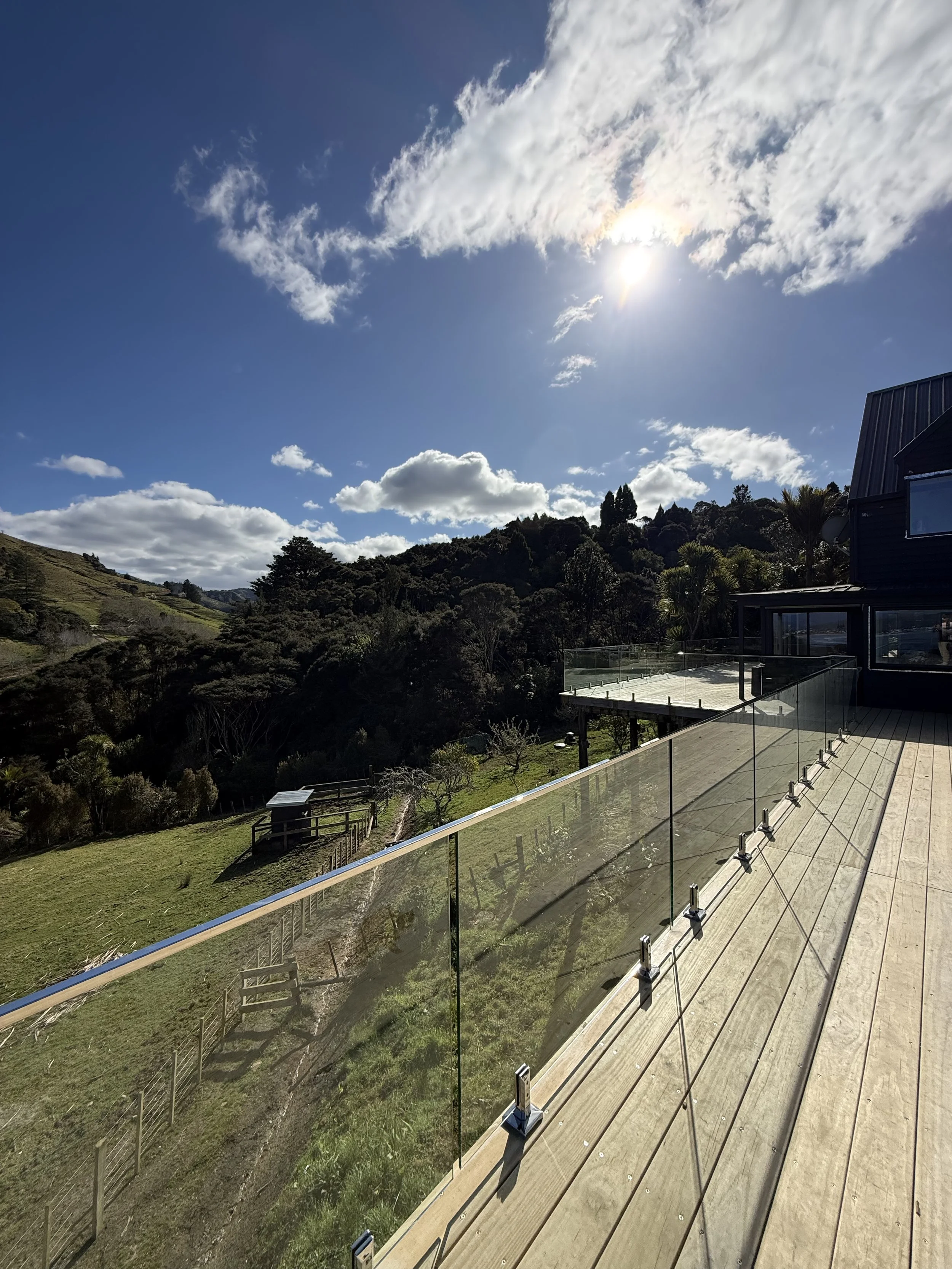 A scenic view of a hillside with trees and grass under a partly cloudy sky with the sun shining brightly, seen from a balcony with glass railings.