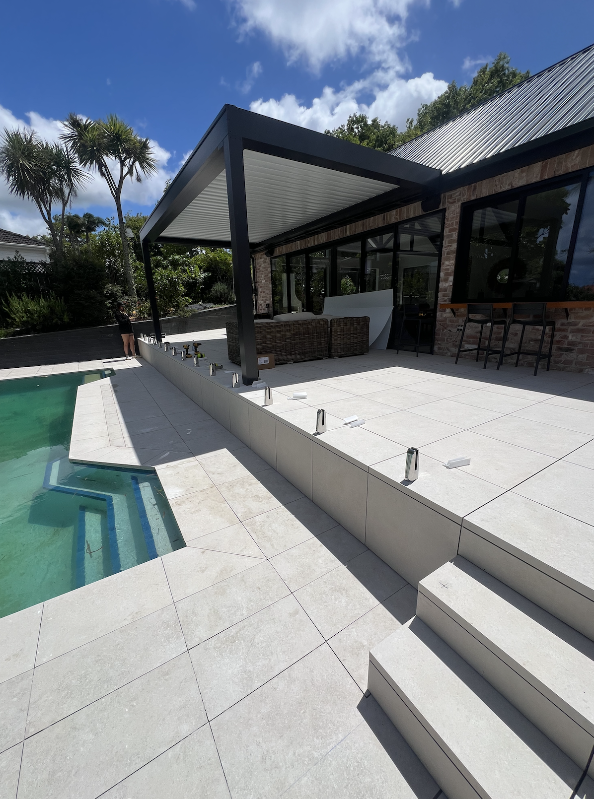 Construction work on a poolside patio with white tiles, stairs, and a covered outdoor seating area with wicker furniture and a brick building.