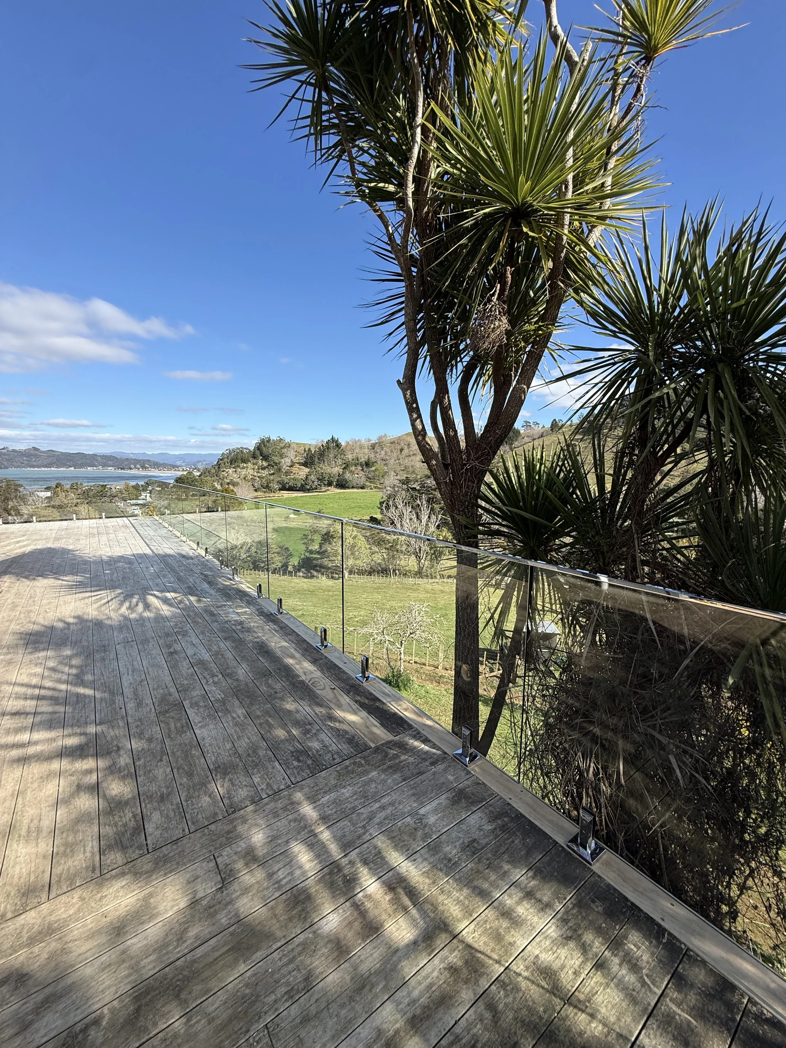 View of a wooden deck with a glass railing overlooking a scenic landscape of green fields, trees, hills, and a river under a partly cloudy blue sky with some white clouds.