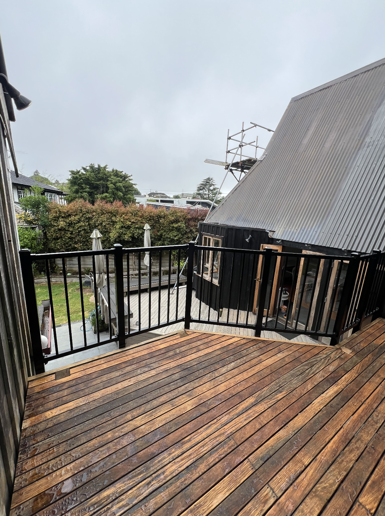 A wooden balcony with a black railing overlooking a backyard with trees, umbrellas, and a neighboring house under overcast sky.