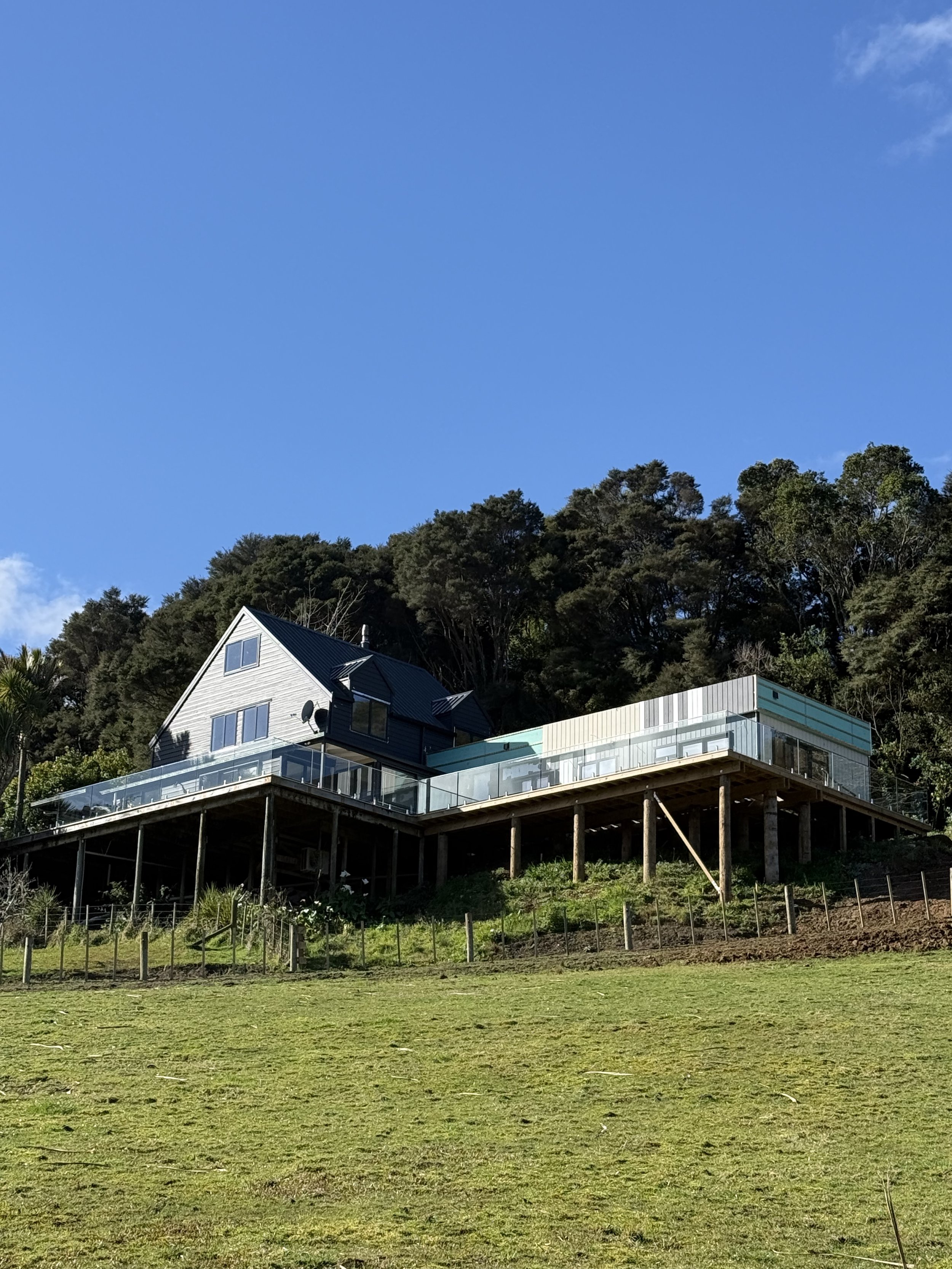 A house elevated on stilts with a deck, located at the edge of a grassy field, with dense trees and a bright blue sky in the background.