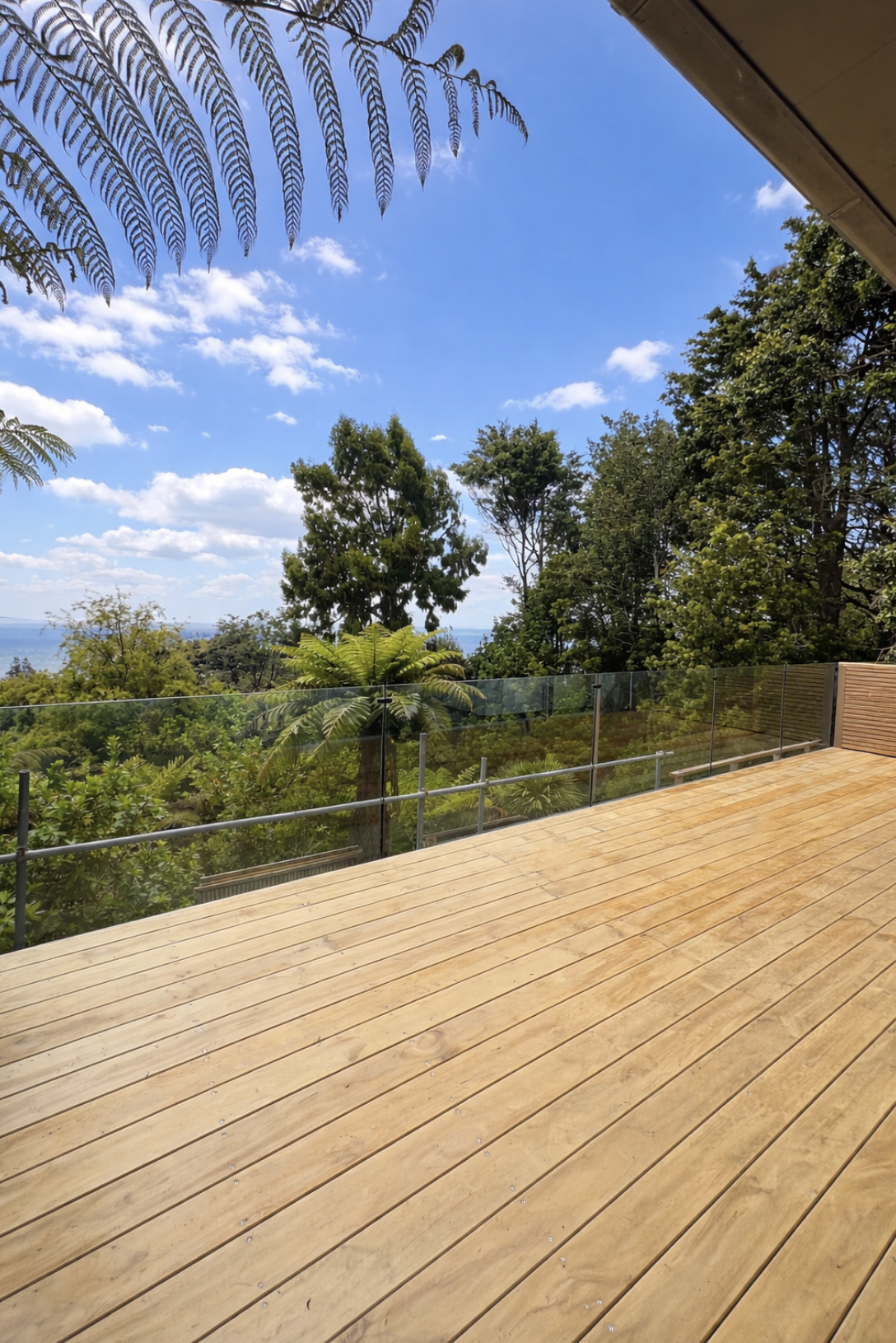 A view of a wooden deck with a glass railing, overlooking a lush green forest with trees and shrubs, under a bright blue sky with scattered clouds.