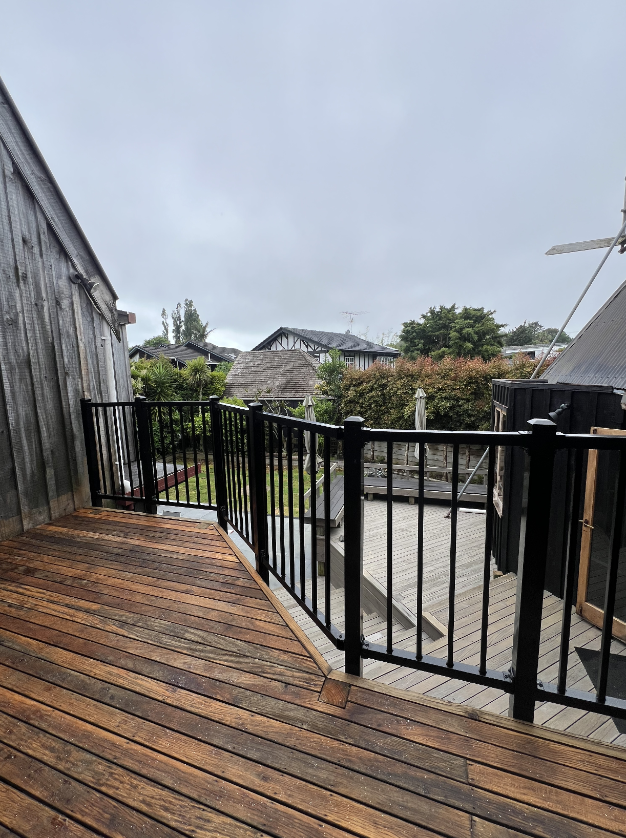 View of a wooden deck with black metal railing overlooking a backyard with trees, shrubs, and neighboring houses under cloudy sky.